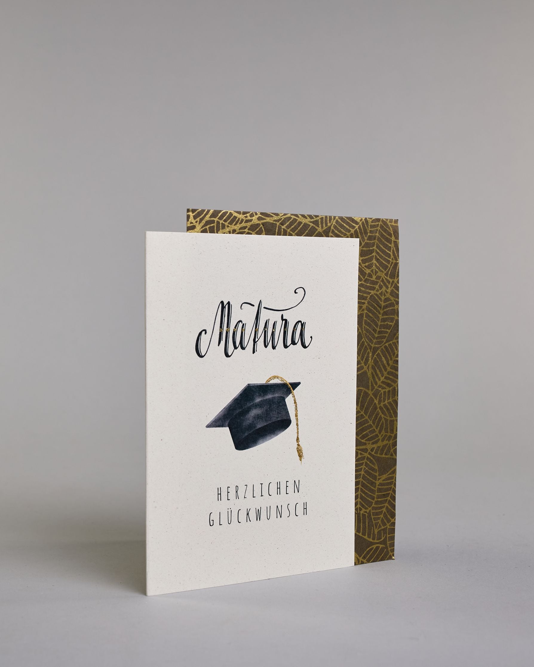 Glückwunschkarte zur Matura mit schwarzer Kalligrafieaufschrift ‚Matura‘, darunter ein schwarzer Doktorhut mit goldener Quaste und der Text ‚Herzlichen Glückwunsch‘ auf hellem Hintergrund. Dazu ein grün-goldenes handgeschöpftes Kuvert. #3