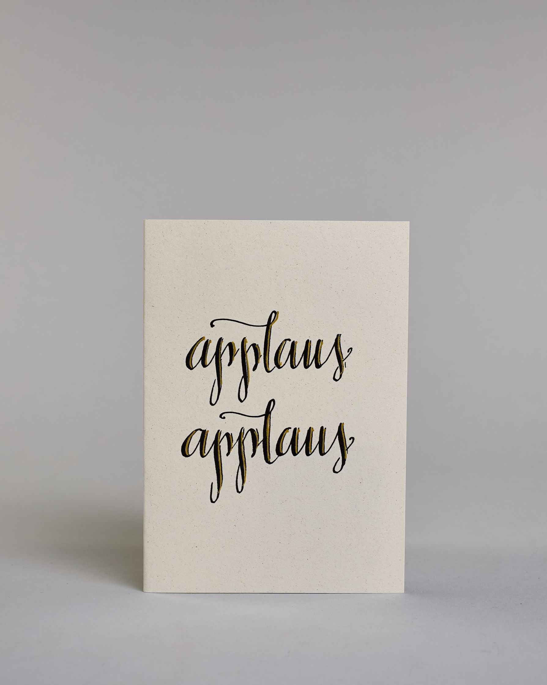 Glückwunschkarte mit schwarzer Kalligrafieaufschrift ‚applaus applaus‘, übereinander geschrieben, Goldverzierung auf hellem Hintergrund. Dazu ein gold-weißes handgeschöpftes Kuvert. #1