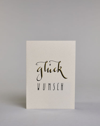Glückwunschkarte mit geschwungener schwarzer Kalligrafieaufschrift ‚glück‘, darunter der Text ‚WUNSCH‘ in geraden Blockbuchstaben auf hellem Hintergrund. Dazu ein weiß-goldenes handgeschöpftes Kuvert. #1