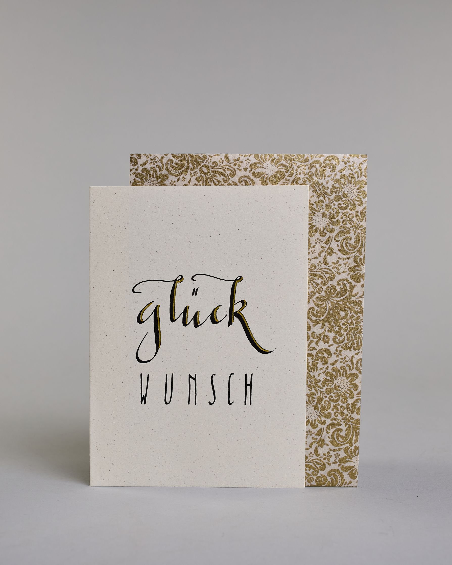 Glückwunschkarte mit geschwungener schwarzer Kalligrafieaufschrift ‚glück‘, darunter der Text ‚WUNSCH‘ in geraden Blockbuchstaben auf hellem Hintergrund. Dazu ein weiß-goldenes handgeschöpftes Kuvert. #2