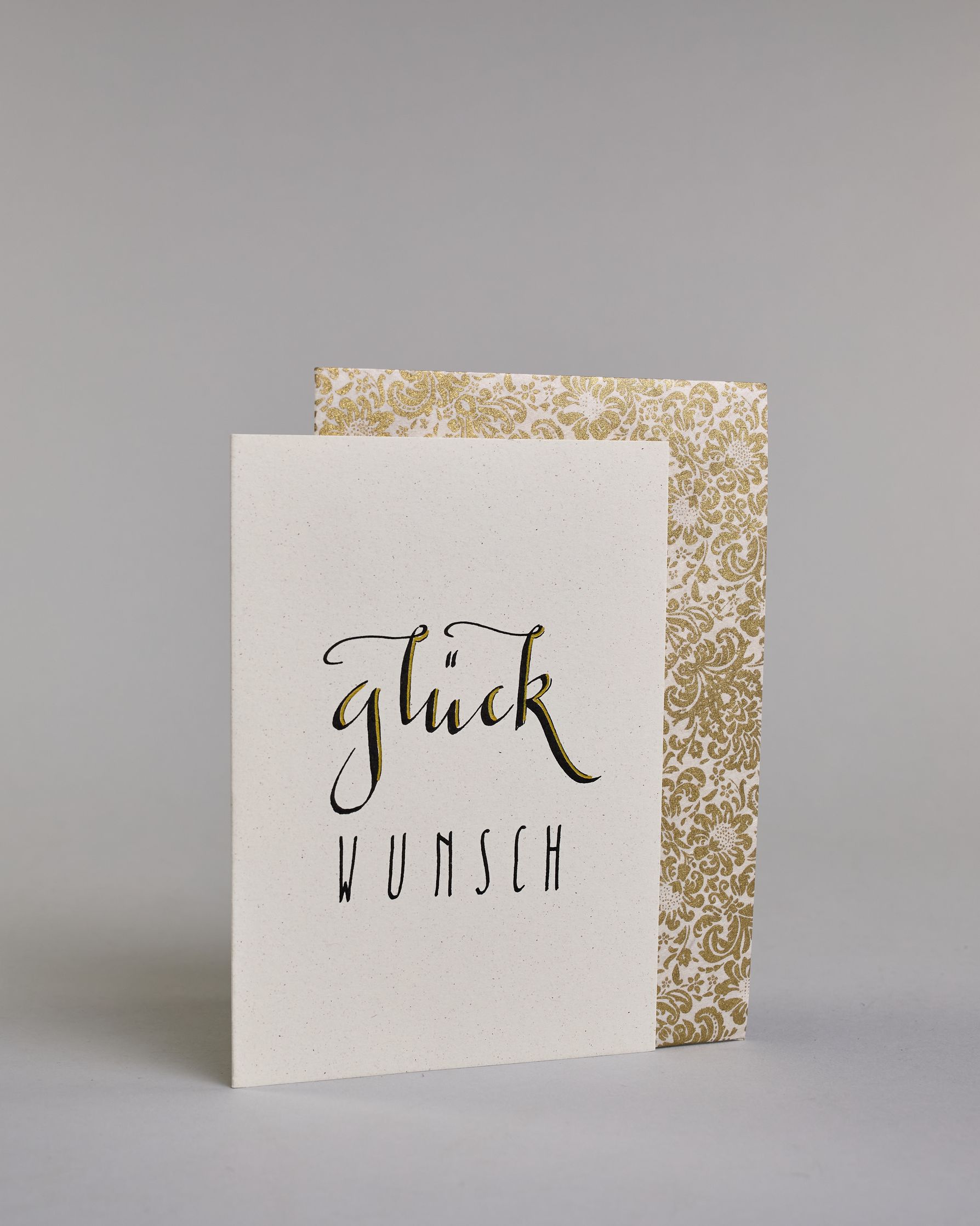 Glückwunschkarte mit geschwungener schwarzer Kalligrafieaufschrift ‚glück‘, darunter der Text ‚WUNSCH‘ in geraden Blockbuchstaben auf hellem Hintergrund. Dazu ein weiß-goldenes handgeschöpftes Kuvert. #3