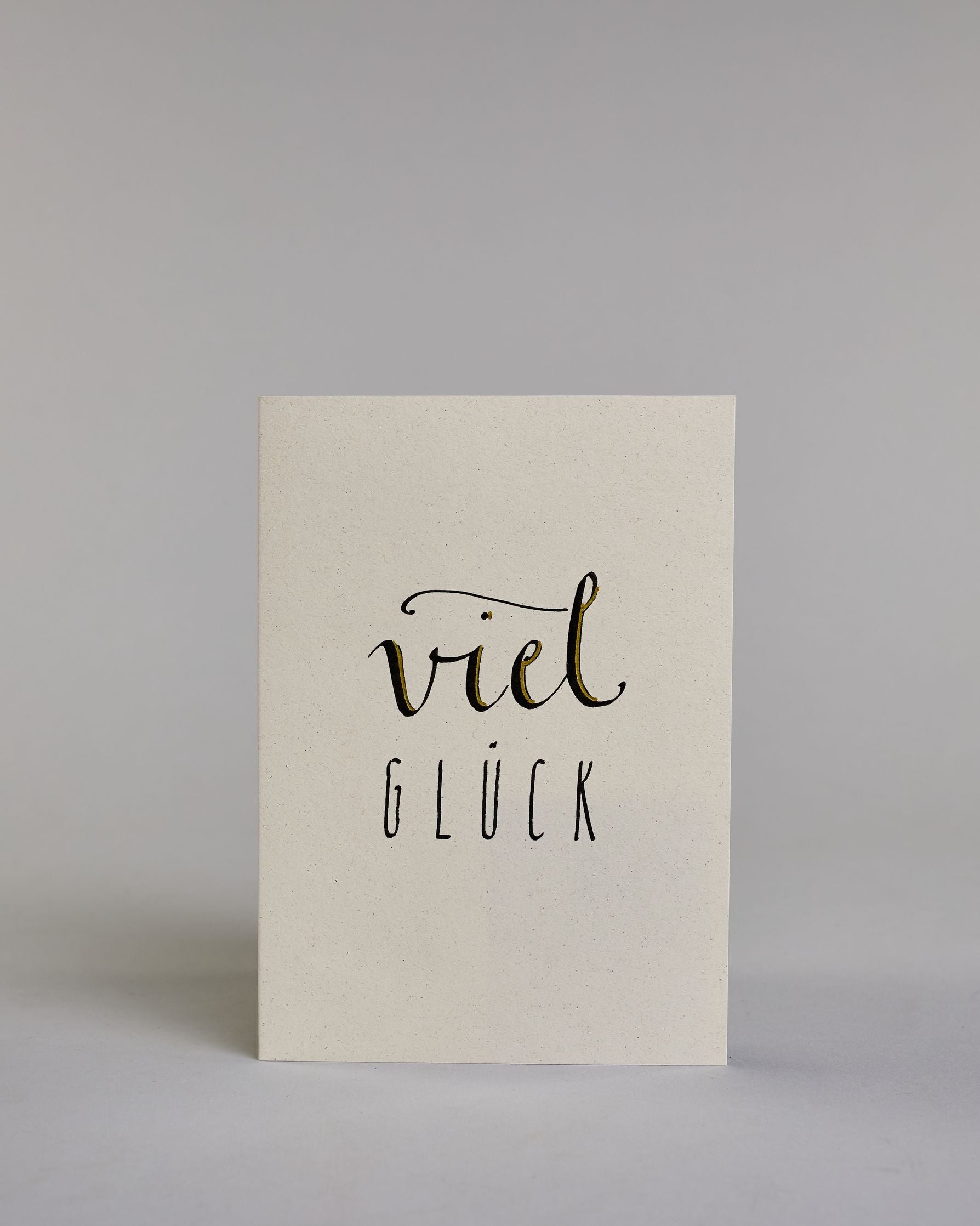 Karte zum viel Erfolg wünschen, mit geschwungener schwarzer Kalligrafieaufschrift ‚viel‘, darunter der Text ‚GLÜCK‘ in geraden Blockbuchstaben auf hellem Hintergrund. Dazu ein grün-goldenes handgeschöpftes Kuvert. #1