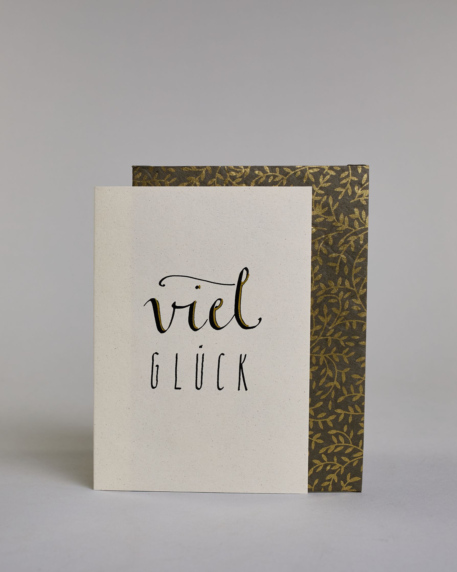 Karte zum viel Erfolg wünschen, mit geschwungener schwarzer Kalligrafieaufschrift ‚viel‘, darunter der Text ‚GLÜCK‘ in geraden Blockbuchstaben auf hellem Hintergrund. Dazu ein grün-goldenes handgeschöpftes Kuvert. #2