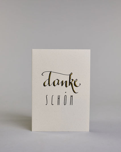 Dankeskarte mit geschwungener schwarzer Kalligrafieaufschrift ‚danke‘, darunter der Text ‚SCHÖN‘ in geraden Blockbuchstaben auf hellem Hintergrund. Dazu ein grün-goldenes handgeschöpftes Kuvert. #1