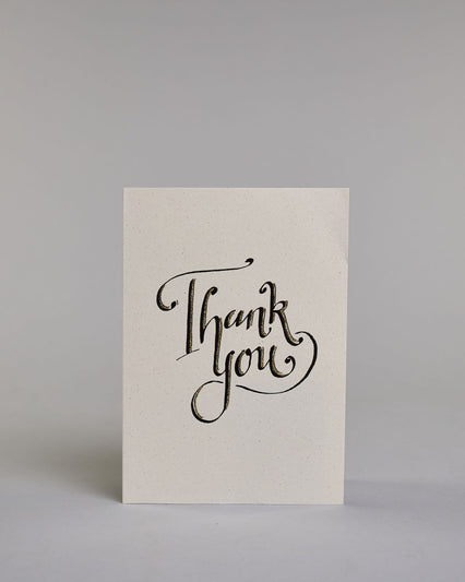 Dankeskarte auf Englisch mit geschwungener schwarzer Kalligrafieaufschrift ‚Thank‘, darunter der Text ‚you‘ auf hellem Hintergrund. Dazu ein grün-goldenes handgeschöpftes Kuvert. #1