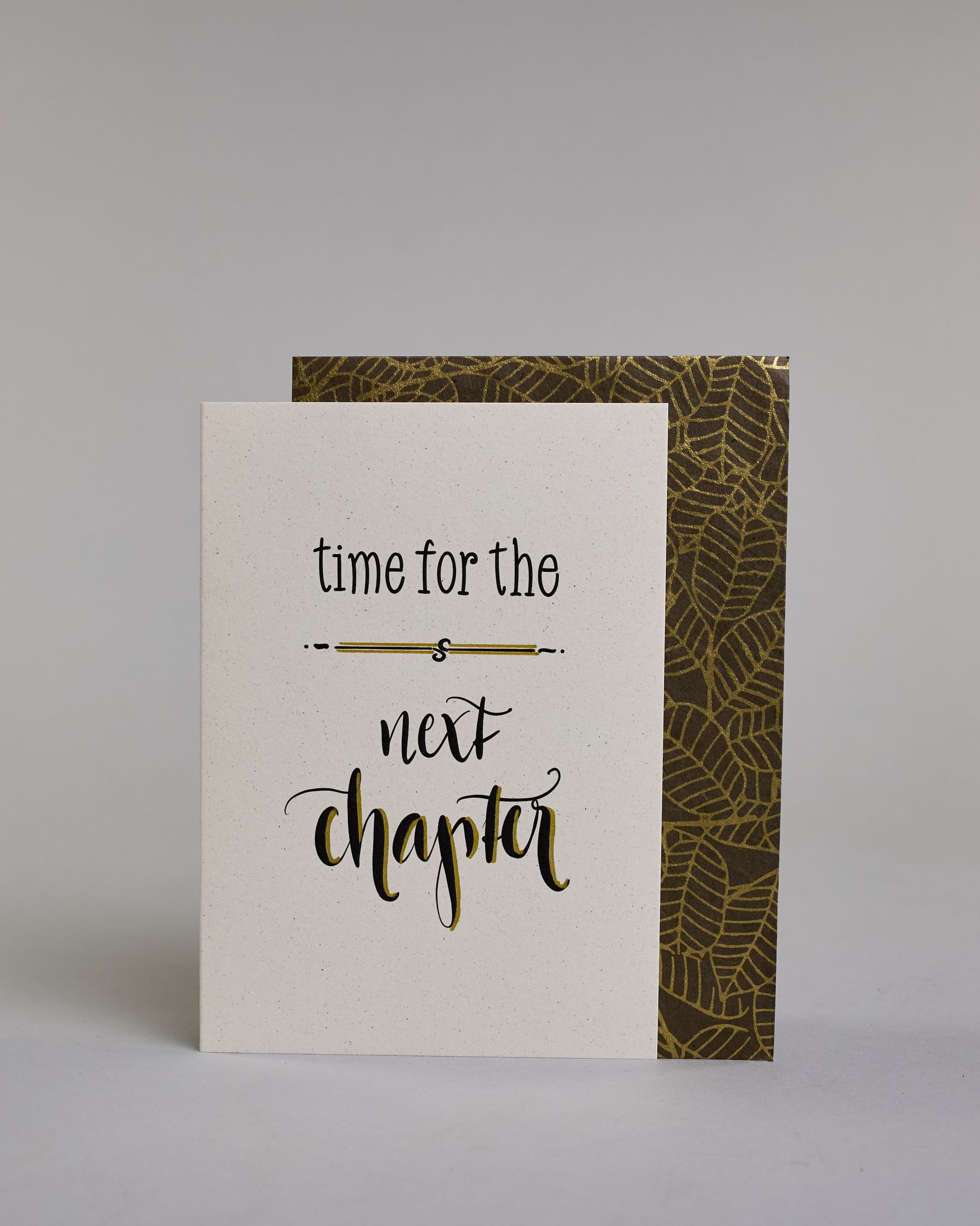 Glückwunschkarte zum Abschied nehmen, auf Englisch mit dem Text ‚time for the next chapter‘. Mit schwarzer Kalligrafieaufschrift ‚time for the‘, darunter eine kunstvolle horizontale Linie, darunter ‚next‘, darunter ‚chapter‘ auf hellem Hintergrund. Dazu ein grün-goldenes Kuvert. #2