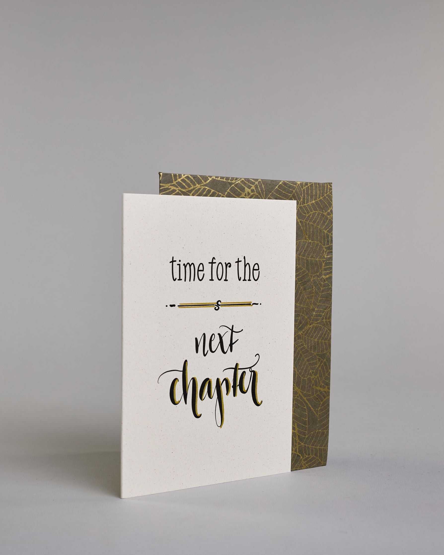 Glückwunschkarte zum Abschied nehmen, auf Englisch mit dem Text ‚time for the next chapter‘. Mit schwarzer Kalligrafieaufschrift ‚time for the‘, darunter eine kunstvolle horizontale Linie, darunter ‚next‘, darunter ‚chapter‘ auf hellem Hintergrund. Dazu ein grün-goldenes Kuvert. #3