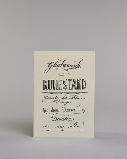 Glückwunschkarte zum Ruhestand, mit dem Text ‚Glückwunsch zum Ruhestand Genieße die schönen Dinge Lebe deine Träume! Danke von uns allen!‘. Aufgeteilt in folgende Zeilen: ‚Glückwunsch‘, darunter ‚zum‘, darunter ‚Ruhestand‘, darunter ‚Genieße die schönen‘, darunter ‚Dinge‘, darunter ‚Lebe deine Träume!‘, darunter ‚Danke‘, darunter ‚von uns allen!‘ auf hellem Hintergrund. Dazu ein grün-goldenes Kuvert. #1