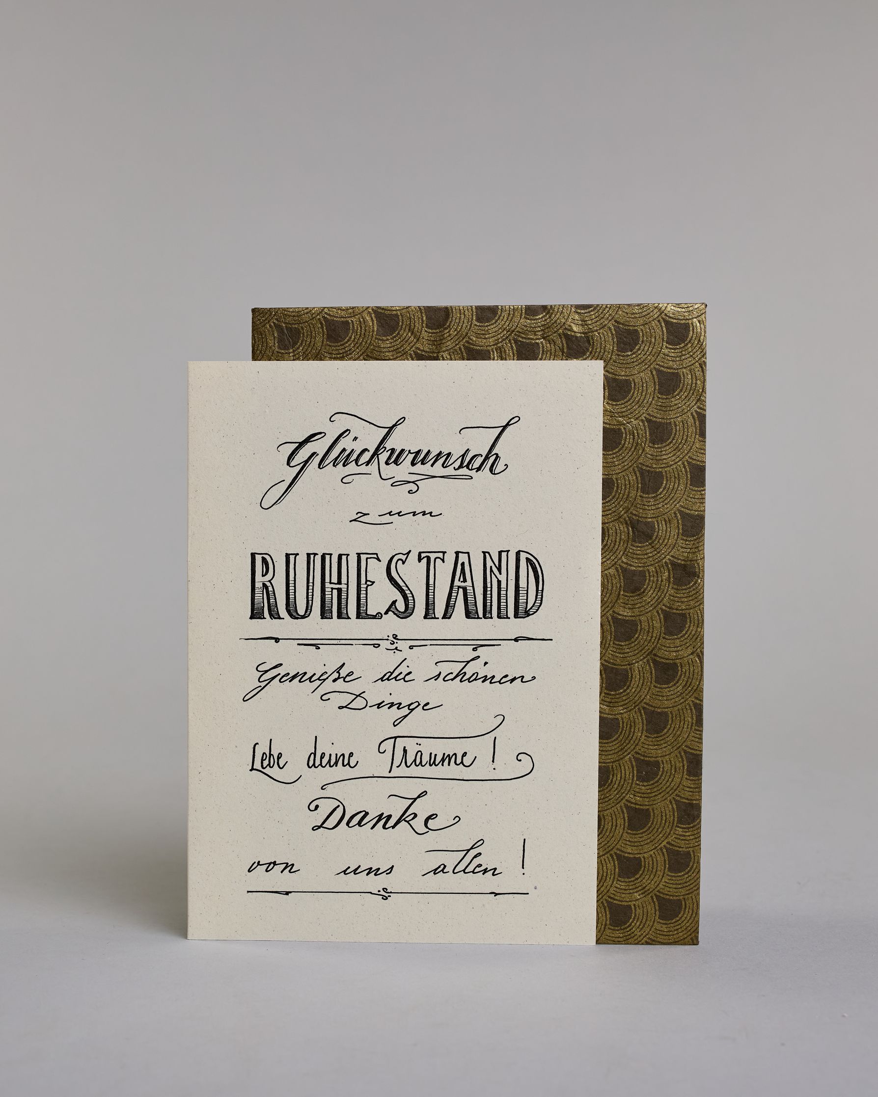 Glückwunschkarte zum Ruhestand, mit dem Text ‚Glückwunsch zum Ruhestand Genieße die schönen Dinge Lebe deine Träume! Danke von uns allen!‘. Aufgeteilt in folgende Zeilen: ‚Glückwunsch‘, darunter ‚zum‘, darunter ‚Ruhestand‘, darunter ‚Genieße die schönen‘, darunter ‚Dinge‘, darunter ‚Lebe deine Träume!‘, darunter ‚Danke‘, darunter ‚von uns allen!‘ auf hellem Hintergrund. Dazu ein grün-goldenes Kuvert. #2