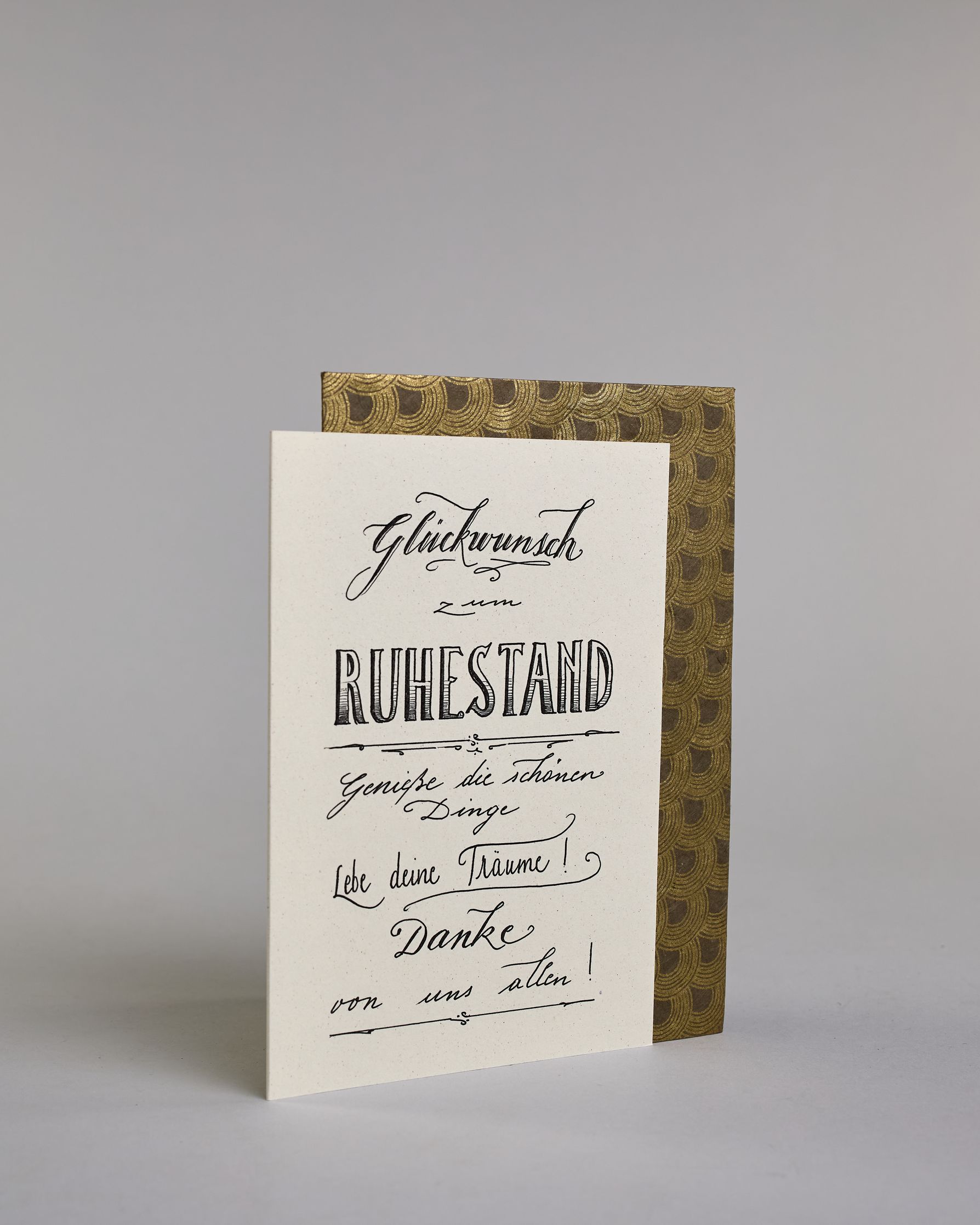 Glückwunschkarte zum Ruhestand, mit dem Text ‚Glückwunsch zum Ruhestand Genieße die schönen Dinge Lebe deine Träume! Danke von uns allen!‘. Aufgeteilt in folgende Zeilen: ‚Glückwunsch‘, darunter ‚zum‘, darunter ‚Ruhestand‘, darunter ‚Genieße die schönen‘, darunter ‚Dinge‘, darunter ‚Lebe deine Träume!‘, darunter ‚Danke‘, darunter ‚von uns allen!‘ auf hellem Hintergrund. Dazu ein grün-goldenes Kuvert. #3