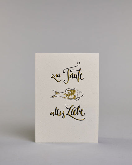 Glückwunschkarte zur Taufe mit geschwungener schwarzer Kalligrafieaufschrift ‚ zur Taufe alles Liebe‘, zwishen den Textzeilen ‚zur Taufe‘ und ‚alles Liebe‘ befindet sich ein gezeichneter goldverzierter Fisch auf hellem Hintergrund. Dazu ein weiß-goldenes handgeschöpftes Kuvert. #1