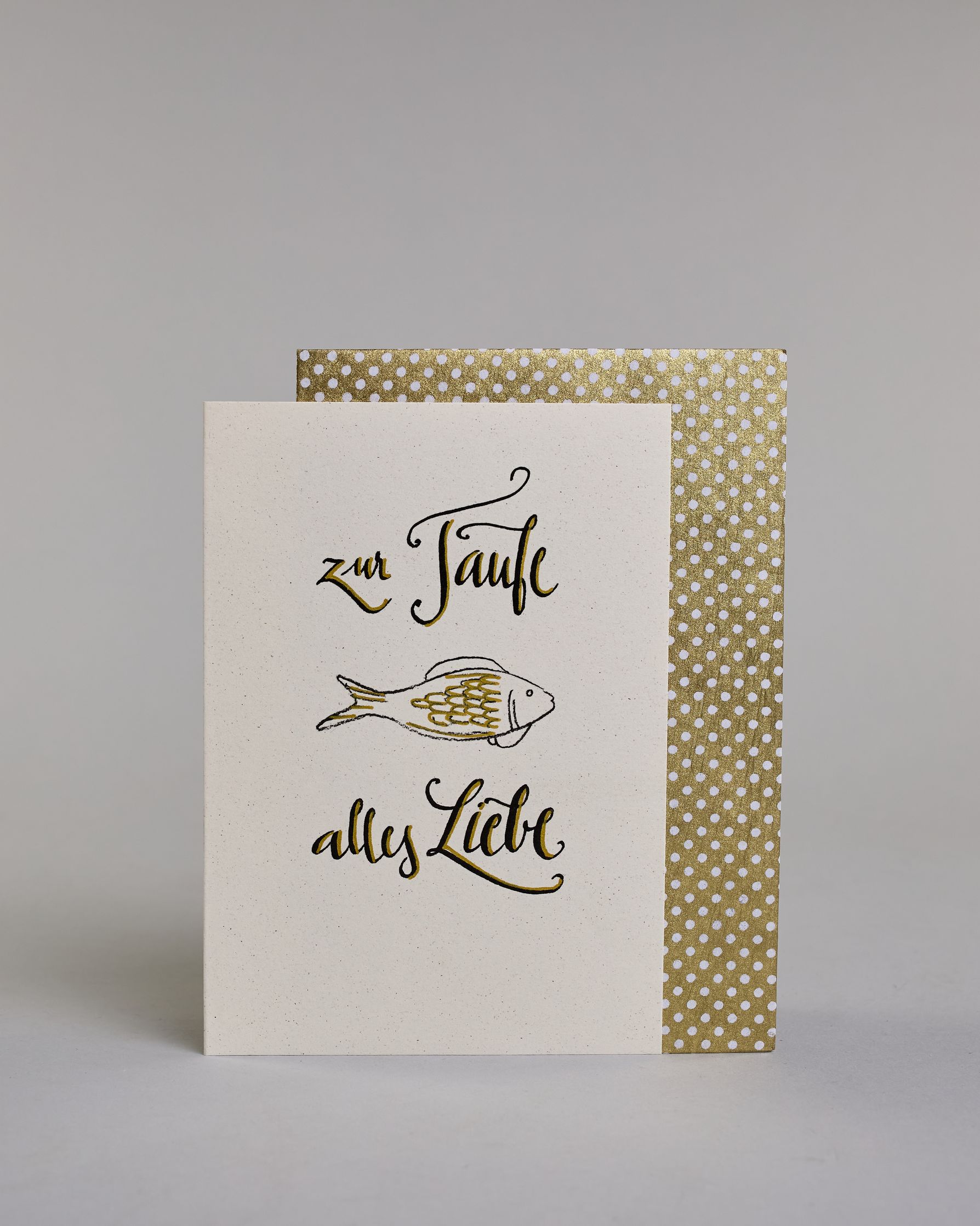 Glückwunschkarte zur Taufe mit geschwungener schwarzer Kalligrafieaufschrift ‚ zur Taufe alles Liebe‘, zwishen den Textzeilen ‚zur Taufe‘ und ‚alles Liebe‘ befindet sich ein gezeichneter goldverzierter Fisch auf hellem Hintergrund. Dazu ein weiß-goldenes handgeschöpftes Kuvert. #2