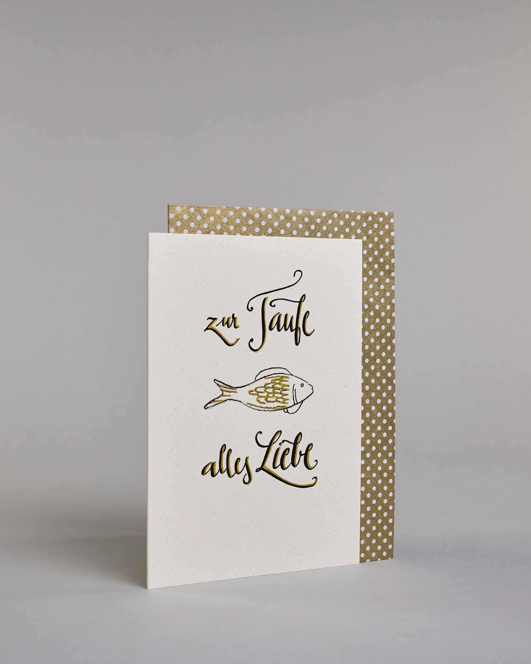 Glückwunschkarte zur Taufe mit geschwungener schwarzer Kalligrafieaufschrift ‚ zur Taufe alles Liebe‘, zwishen den Textzeilen ‚zur Taufe‘ und ‚alles Liebe‘ befindet sich ein gezeichneter goldverzierter Fisch auf hellem Hintergrund. Dazu ein weiß-goldenes handgeschöpftes Kuvert. #3