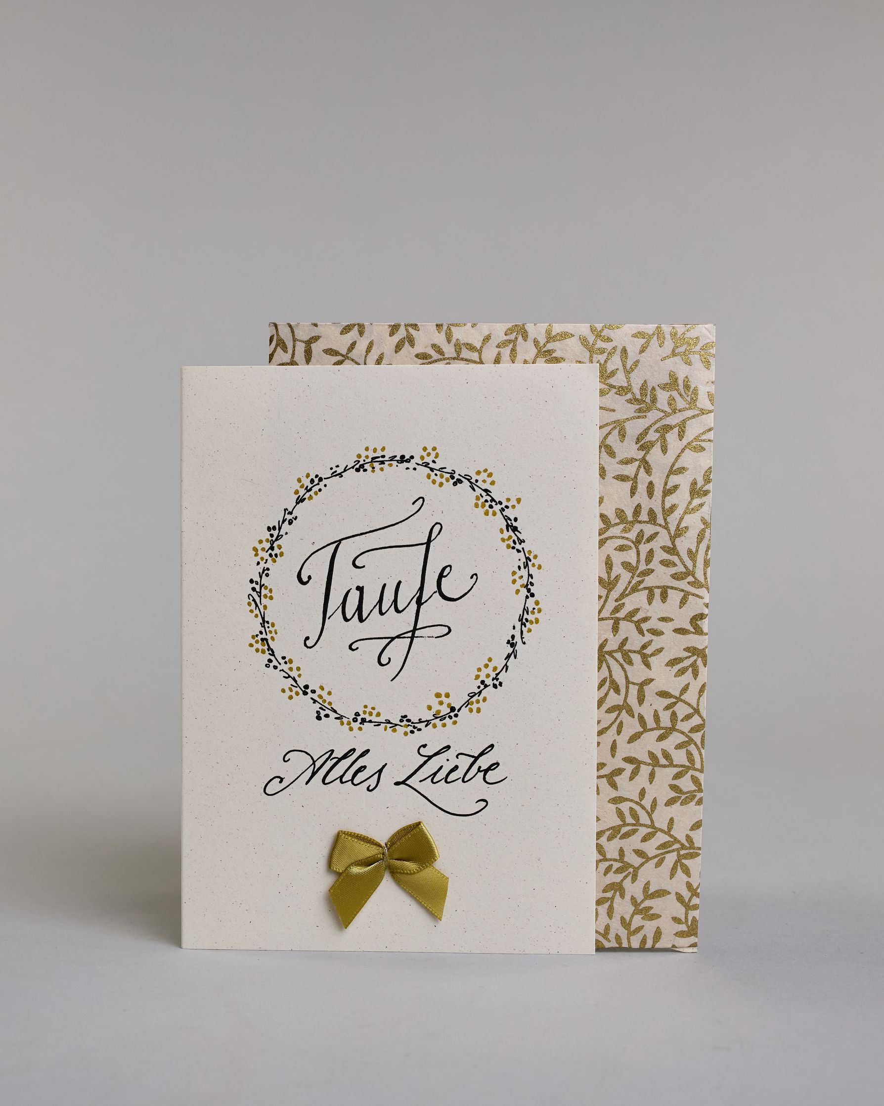 Glückwunschkarte zur Taufe mit geschwungener schwarzer Kalligrafieaufschrift in gezeichnetem Kranz ‚Taufe‘, darunter der Text ‚Alles Liebe‘, darunter eine aufgenähte goldene Masche auf hellem Hintergrund. Dazu ein weiß-goldenes handgeschöpftes Kuvert. #2