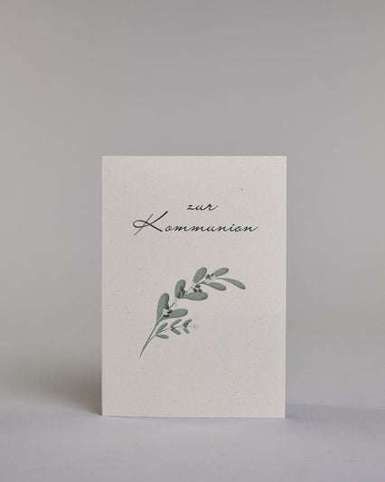 Glückwunschkarte zur Kommunion mit geschwungener schwarzer Kalligrafieaufschrift ‚zur Kommunion‘, darunter ein grüner Zweig mit Oliven und weißen Blüten auf hellem Hintergrund. Dazu ein blau-weißes handgeschöpftes Kuvert. #1