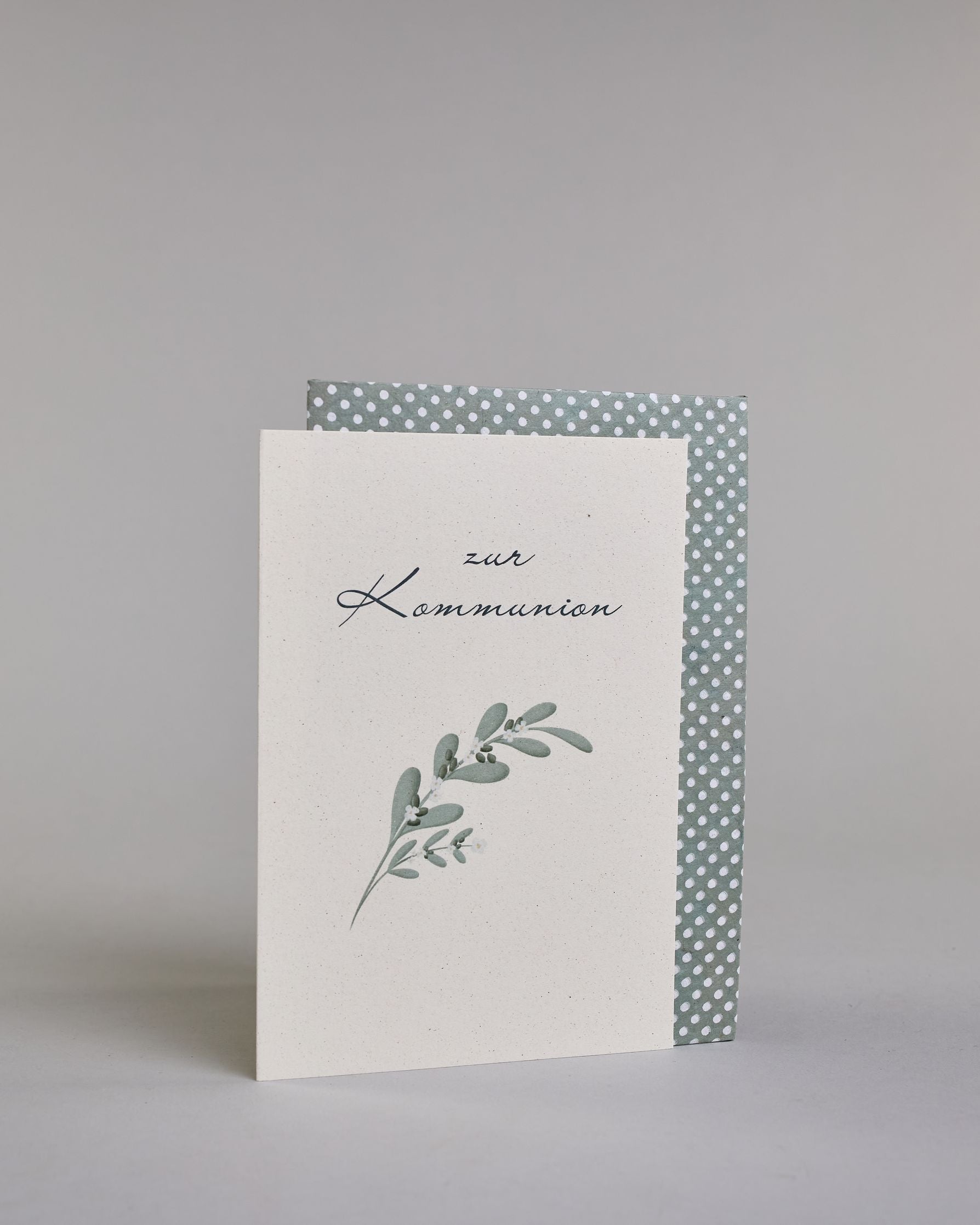 Glückwunschkarte zur Kommunion mit geschwungener schwarzer Kalligrafieaufschrift ‚zur Kommunion‘, darunter ein grüner Zweig mit Oliven und weißen Blüten auf hellem Hintergrund. Dazu ein blau-weißes handgeschöpftes Kuvert. #3