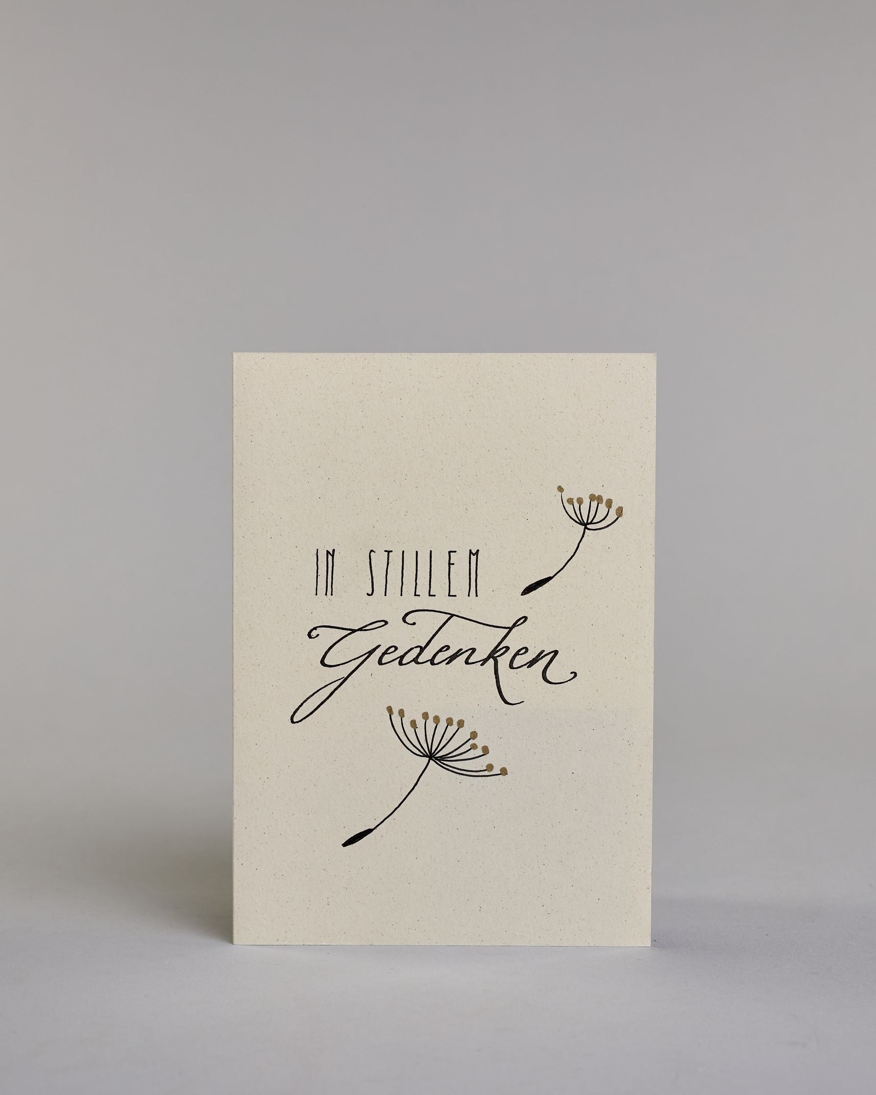 Trauerkarte zur Kondolenzbekundung mit schwarzer Kalligrafieaufschrift ‚in stillem Gedenken‘,  ‚IN STILLEM ‘ mit gerader schwarzer Kalligrafieaufschrift daneben ein goldverzierter einzelner Strang einer Pusteblume‚ darunter ‚Gedenken‘ in geschwungener schwarzer Kalligrafieaufschrift, darunter ein weiterer goldverzierter Strang einer Pusteblume auf hellem Hintergrund. Dazu ein schwarzes handgeschöpftes Kuvert. #1