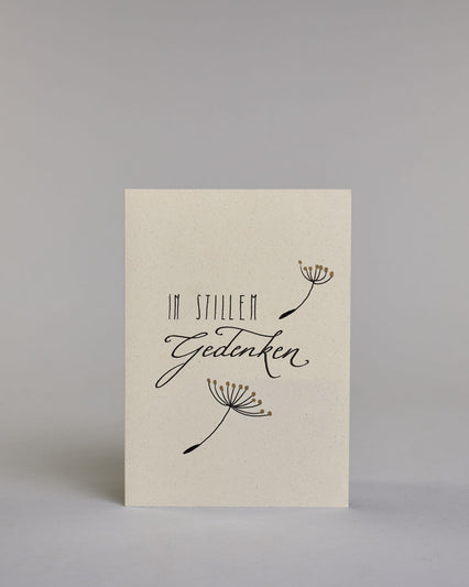 Trauerkarte zur Kondolenzbekundung mit schwarzer Kalligrafieaufschrift ‚in stillem Gedenken‘,  ‚IN STILLEM ‘ mit gerader schwarzer Kalligrafieaufschrift daneben ein goldverzierter einzelner Strang einer Pusteblume‚ darunter ‚Gedenken‘ in geschwungener schwarzer Kalligrafieaufschrift, darunter ein weiterer goldverzierter Strang einer Pusteblume auf hellem Hintergrund. Dazu ein schwarzes handgeschöpftes Kuvert. #1