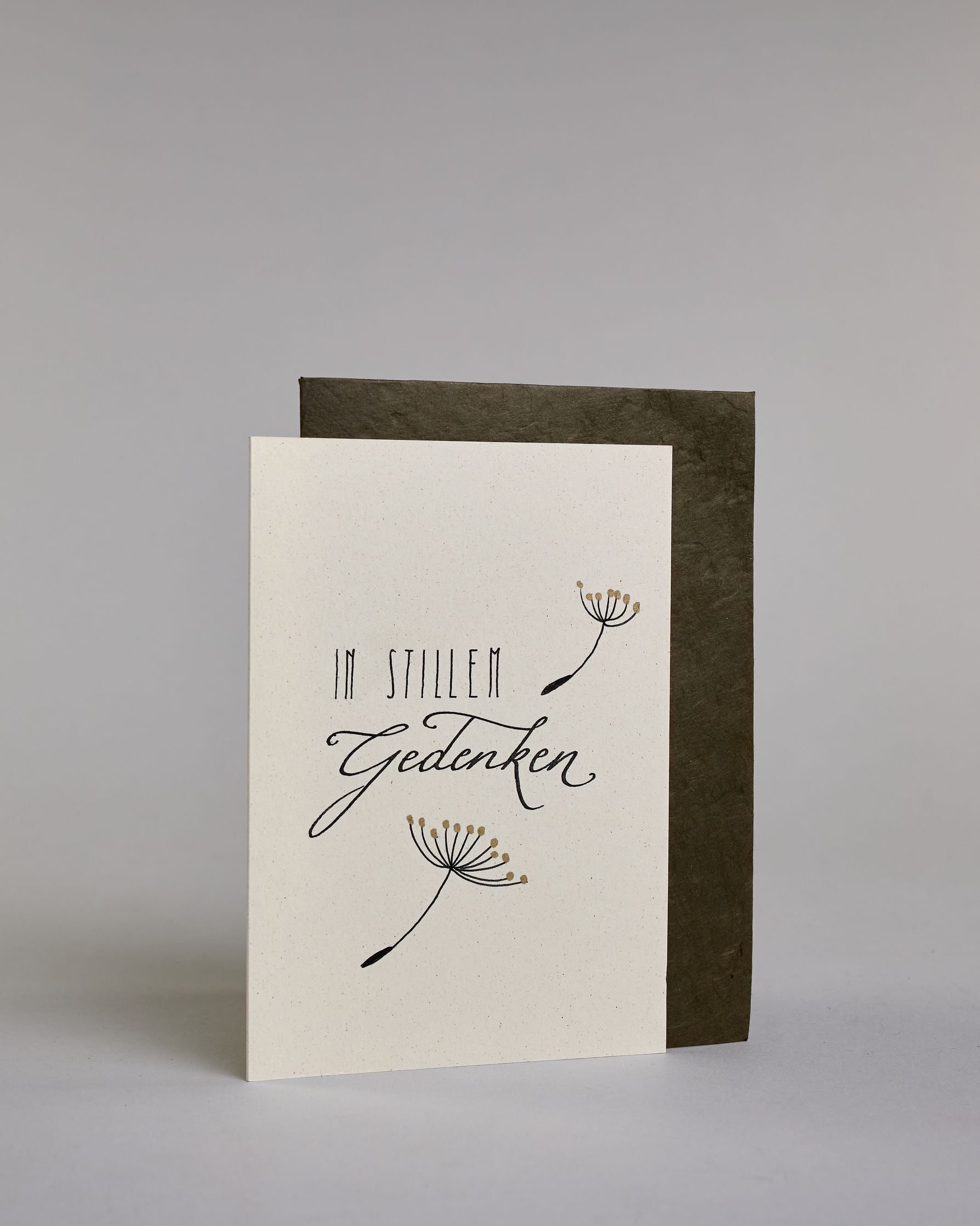 Trauerkarte zur Kondolenzbekundung mit schwarzer Kalligrafieaufschrift ‚in stillem Gedenken‘,  ‚IN STILLEM ‘ mit gerader schwarzer Kalligrafieaufschrift daneben ein goldverzierter einzelner Strang einer Pusteblume‚ darunter ‚Gedenken‘ in geschwungener schwarzer Kalligrafieaufschrift, darunter ein weiterer goldverzierter Strang einer Pusteblume auf hellem Hintergrund. Dazu ein schwarzes handgeschöpftes Kuvert. #3