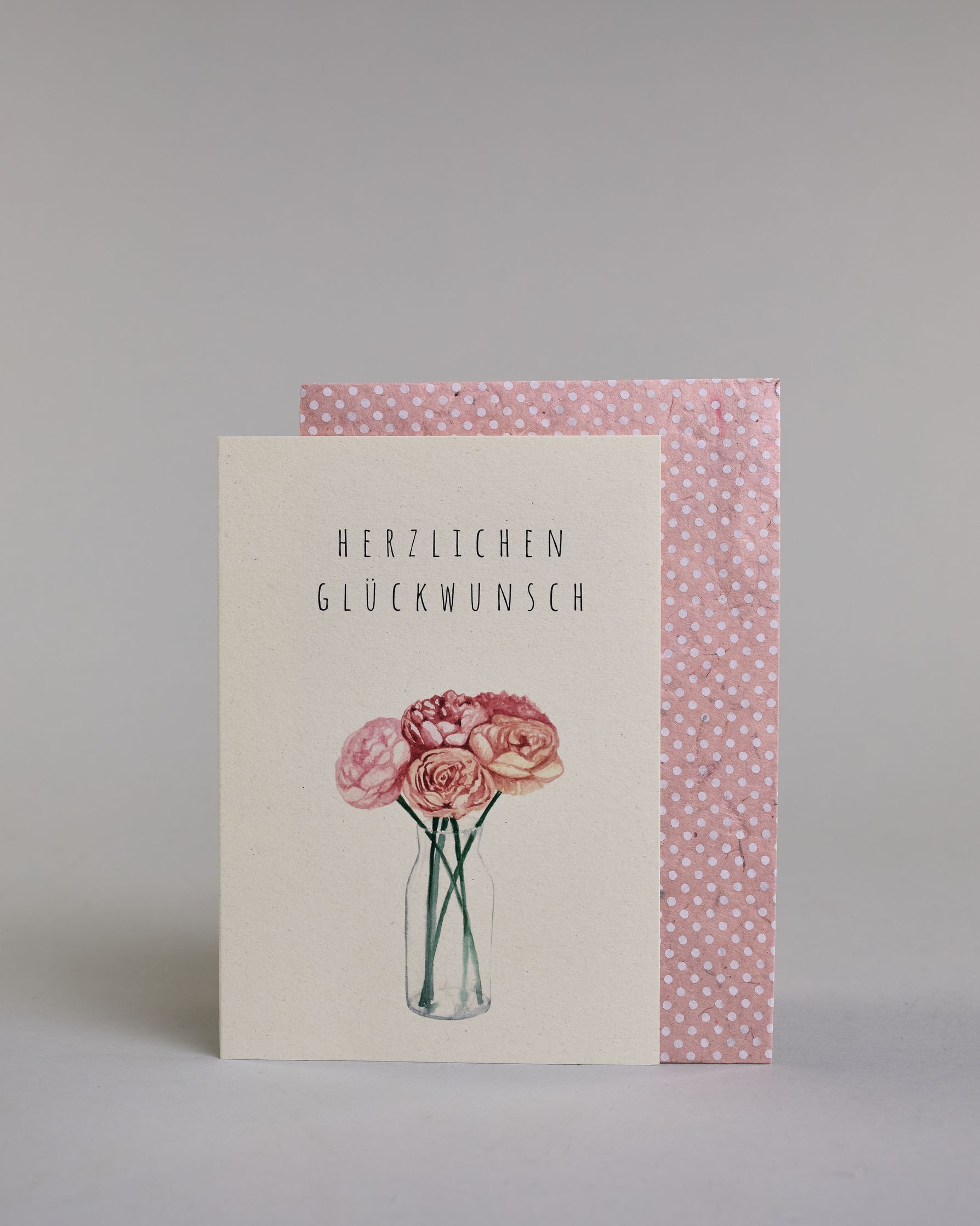 Allgemeine Glückwunschkarte mit schwarzer Kalligrafieaufschrift ‚Herzlichen Glückwunsch‘, darunter ist ein gezeichneter Blumenstrauß mit rosa Blüten in einer Vase auf hellem Hintergrund. Dazu ein rosa-weißes handgeschöpftes Kuvert. #2