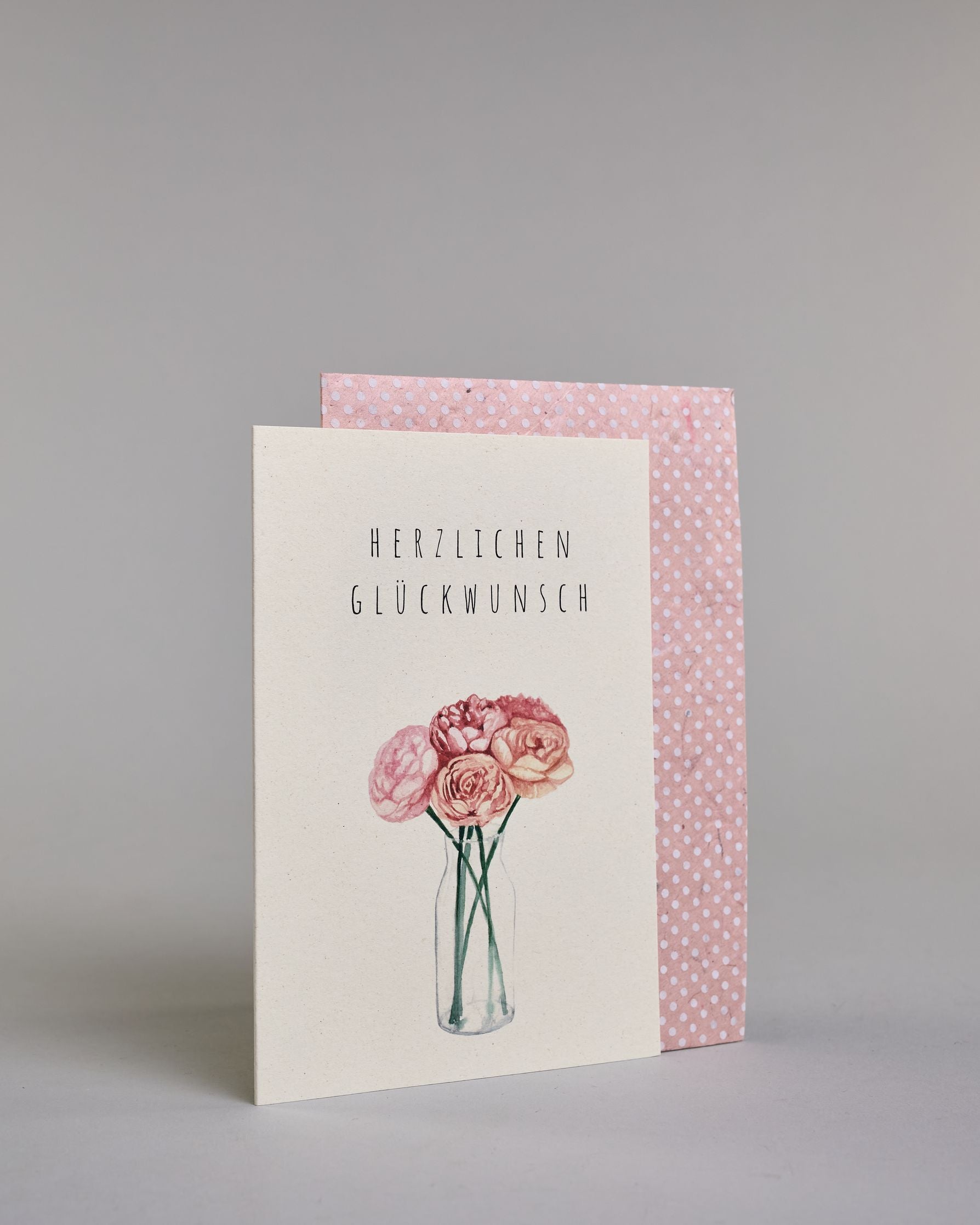 Allgemeine Glückwunschkarte mit schwarzer Kalligrafieaufschrift ‚Herzlichen Glückwunsch‘, darunter ist ein gezeichneter Blumenstrauß mit rosa Blüten in einer Vase auf hellem Hintergrund. Dazu ein rosa-weißes handgeschöpftes Kuvert. #3