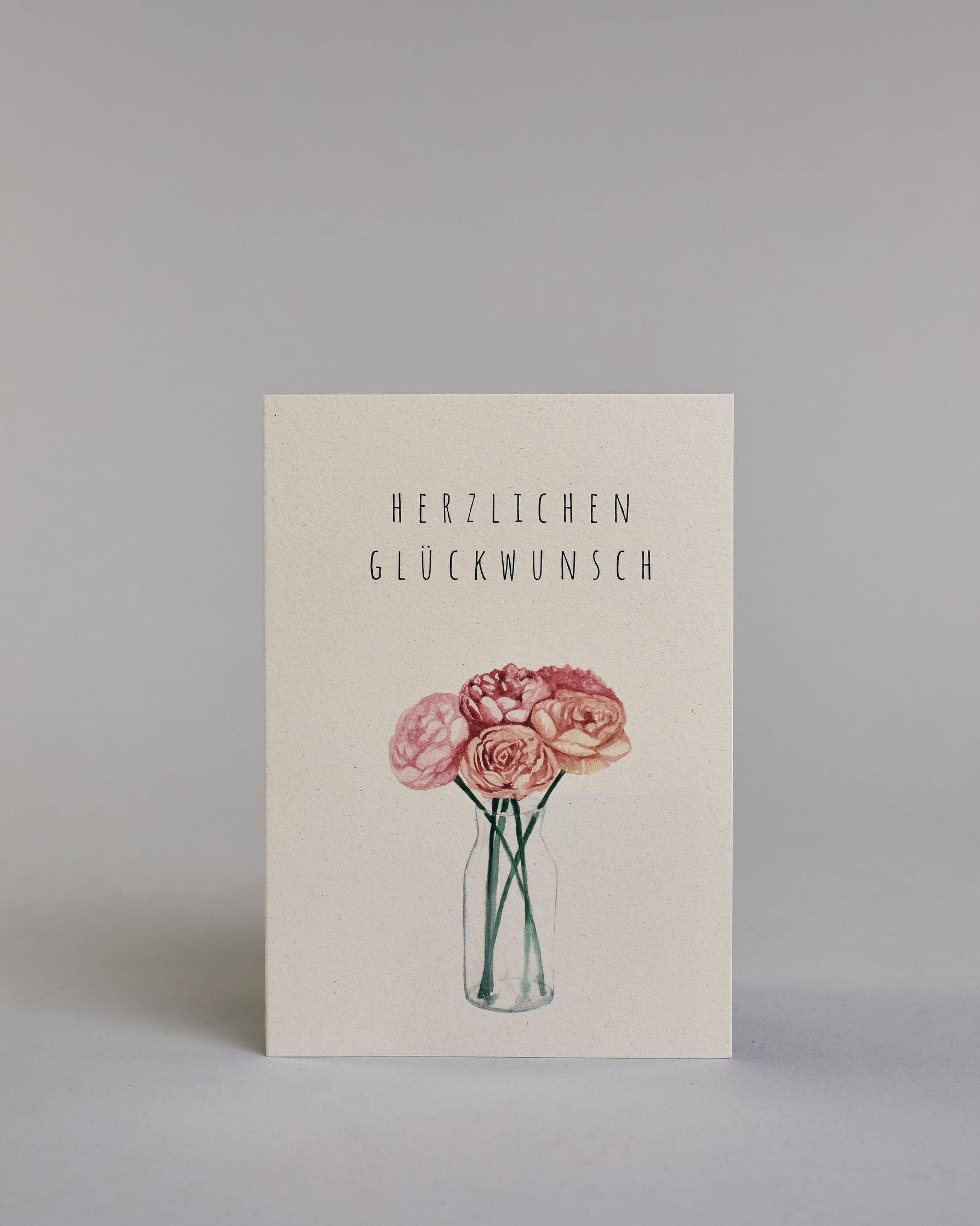Allgemeine Glückwunschkarte mit schwarzer Kalligrafieaufschrift ‚Herzlichen Glückwunsch‘, darunter ist ein gezeichneter Blumenstrauß mit rosa Blüten in einer Vase auf hellem Hintergrund. Dazu ein rosa-weißes handgeschöpftes Kuvert. #1