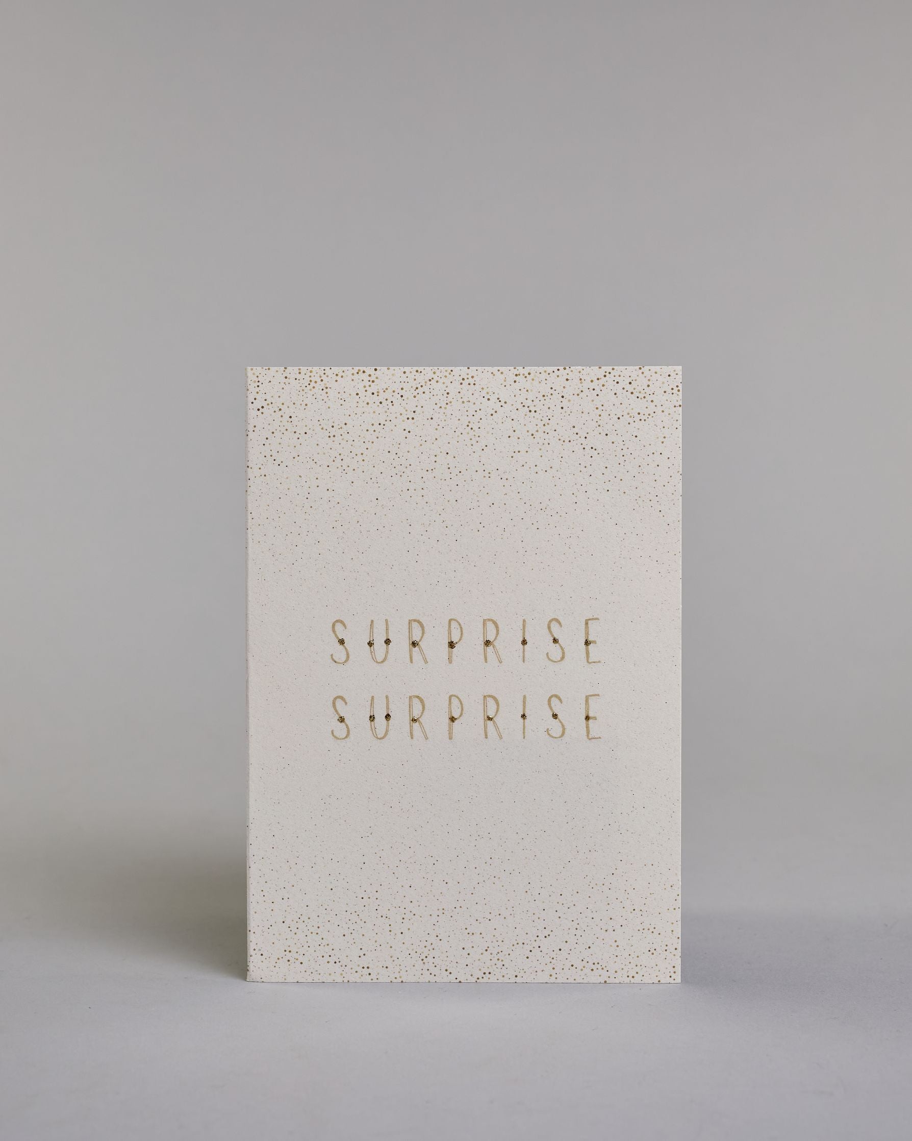 Karte in Englisch mit goldener Kalligrafieaufschrift ‚surprise surprise‘, von der oberen und unteren Kartenkante kommt ein Pünktchenregen auf hellem Hintergrund. Dazu ein weißes handgeschöpftes Kuvert. #1