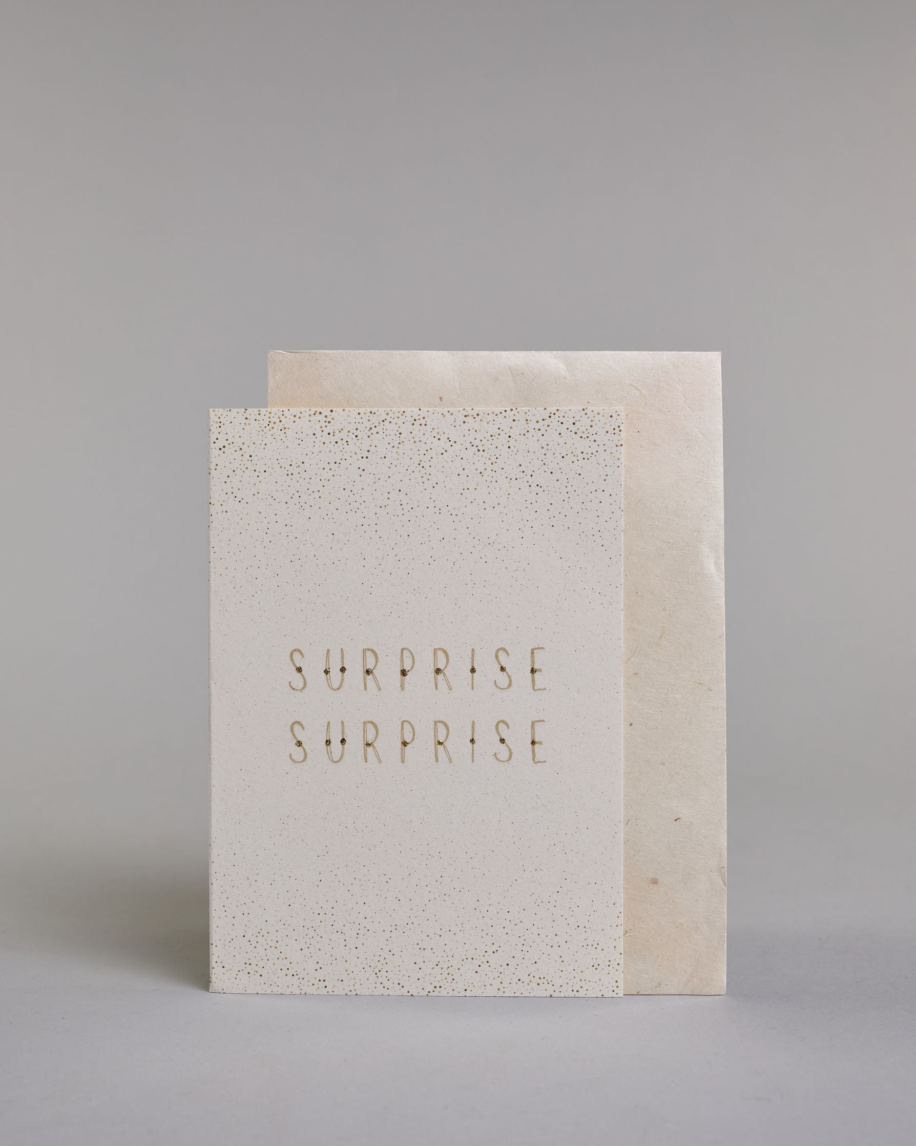 Karte in Englisch mit goldener Kalligrafieaufschrift ‚surprise surprise‘, von der oberen und unteren Kartenkante kommt ein Pünktchenregen auf hellem Hintergrund. Dazu ein weißes handgeschöpftes Kuvert. #2