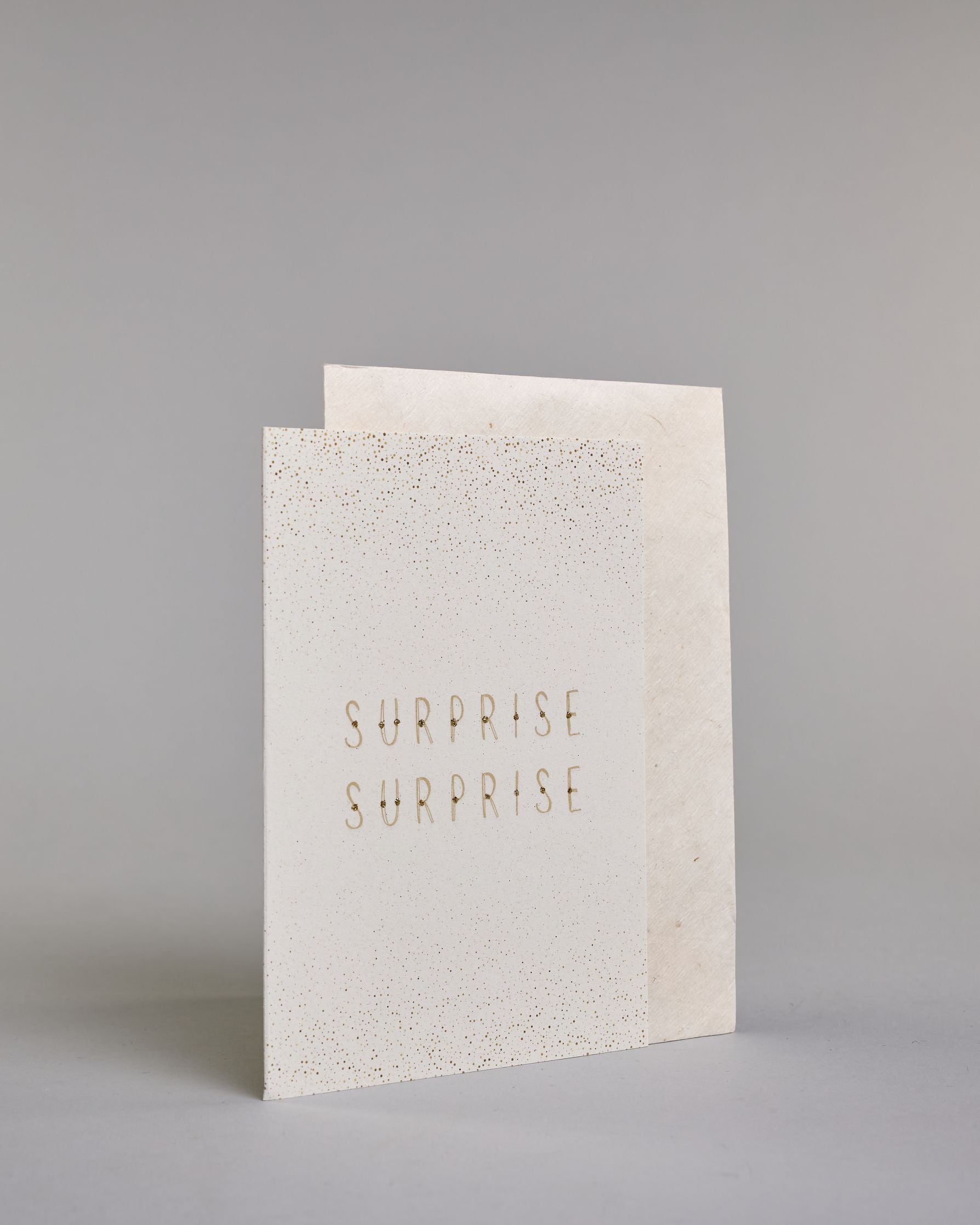 Karte in Englisch mit goldener Kalligrafieaufschrift ‚surprise surprise‘, von der oberen und unteren Kartenkante kommt ein Pünktchenregen auf hellem Hintergrund. Dazu ein weißes handgeschöpftes Kuvert. #3