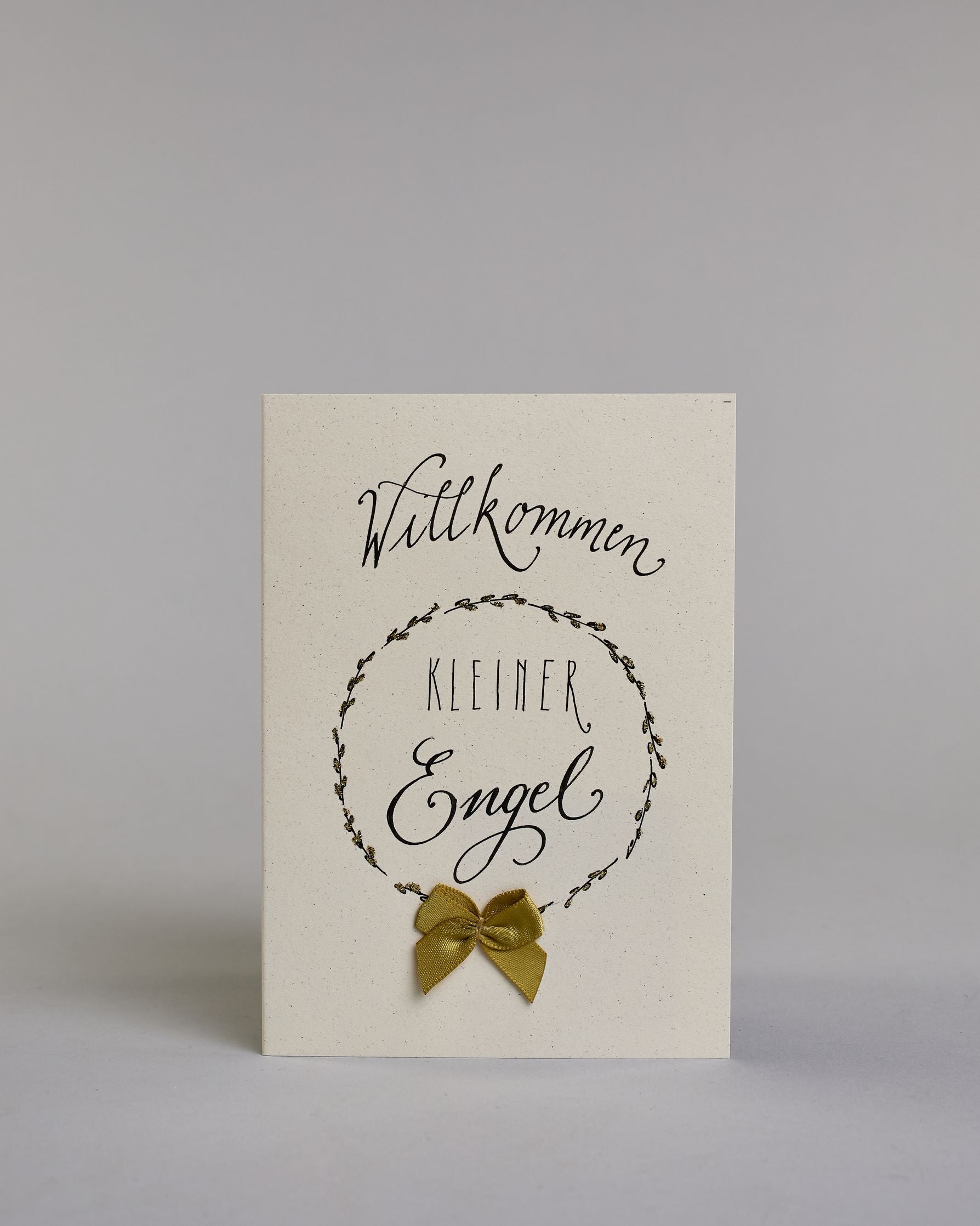 Glückwunschkarte zur Geburt mit schwarzer Kalligrafieaufschrift ‚Willkommen kleiner Engel‘, ‚Willkommen‘ steht in geschwungener Kalligrafieaufschrift über einem gezeichnetem Kranz, untem auf dem Kranz ist eine goldene Masche aufgenäht. Der Text im Kranz setzt sich auf folgenden Zeilen zusammen:  ‚KLEINER‘ in dünner gerader Kalligrafieaufschrift, darunter ‚Engel‘, auf hellem Hintergrund. Dazu ein gold-weißes handgeschöpftes Kuvert. #1