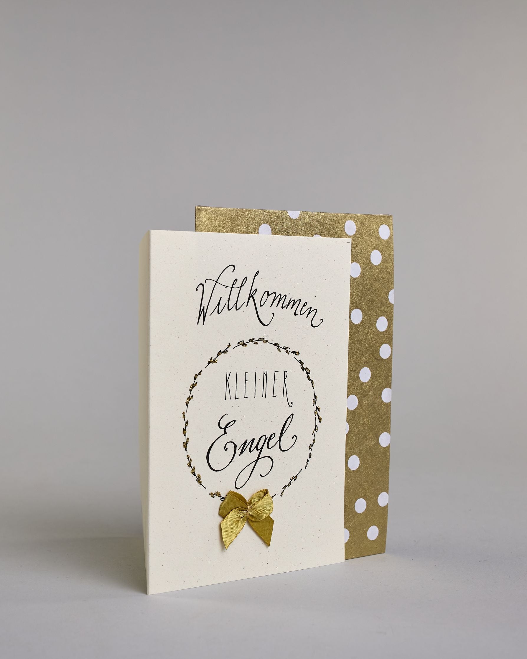 Glückwunschkarte zur Geburt mit schwarzer Kalligrafieaufschrift ‚Willkommen kleiner Engel‘, ‚Willkommen‘ steht in geschwungener Kalligrafieaufschrift über einem gezeichnetem Kranz, untem auf dem Kranz ist eine goldene Masche aufgenäht. Der Text im Kranz setzt sich auf folgenden Zeilen zusammen:  ‚KLEINER‘ in dünner gerader Kalligrafieaufschrift, darunter ‚Engel‘, auf hellem Hintergrund. Dazu ein gold-weißes handgeschöpftes Kuvert. #3