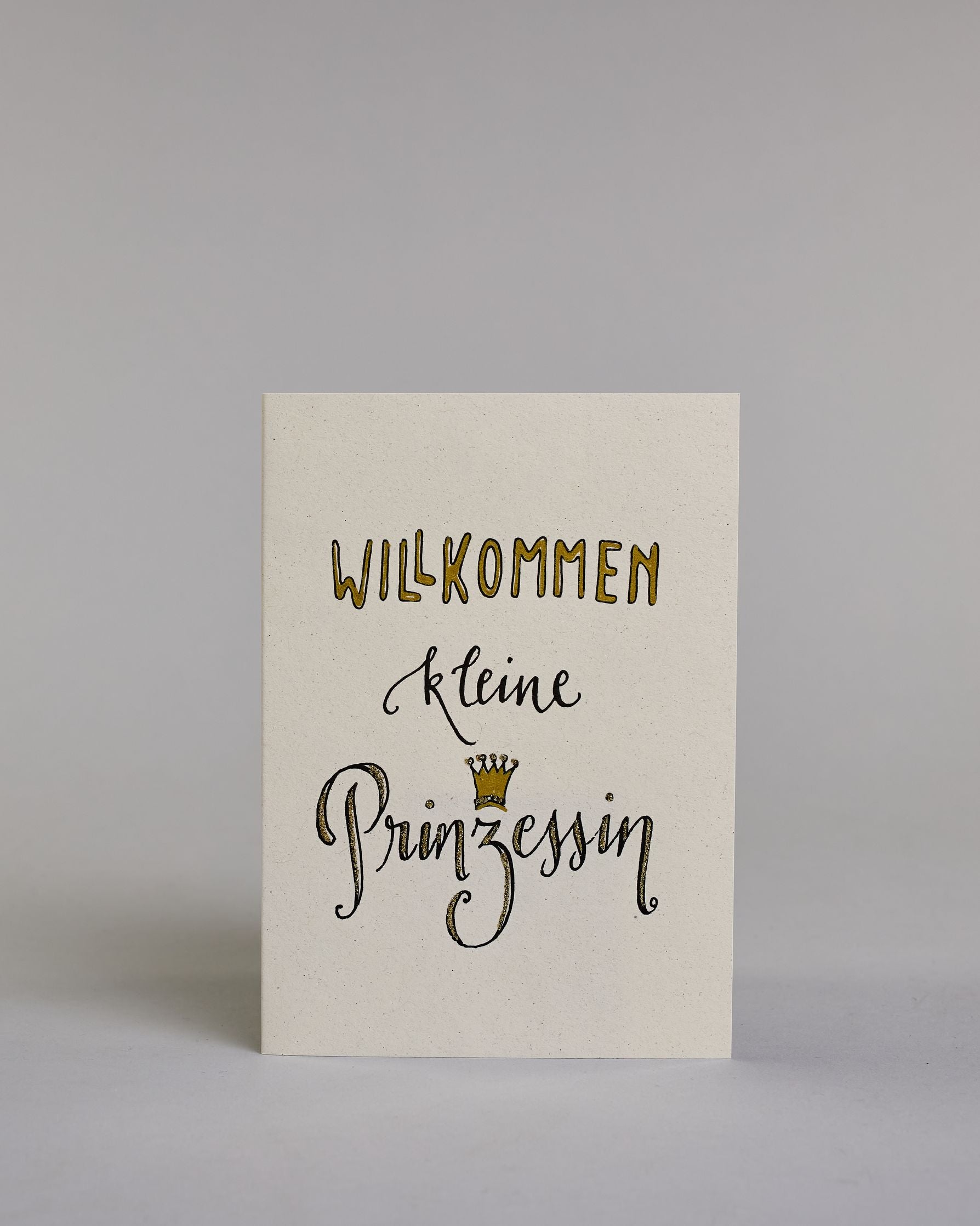 Glückwunschkarte zur Geburt mit schwarzer Kalligrafieaufschrift ‚Willkommen kleine Prinzessin‘, der Text setzt sich auf folgenden Zeilen zusammen:  ‚WILLKOMMEN‘ in gold gefüllten Kalligrafiebuchstaben, darunter‚kleine‘ in geschwungener schwarzer Kalligrafieaufschrift, darunter ‚Prinzessin‘ in verglitzerter Kalligrafieaufschrift, über dem ‚z‘ ist eine gezeichnete goldene Krone auf hellem Hintergrund. Dazu ein gold-weißes handgeschöpftes Kuvert. #1