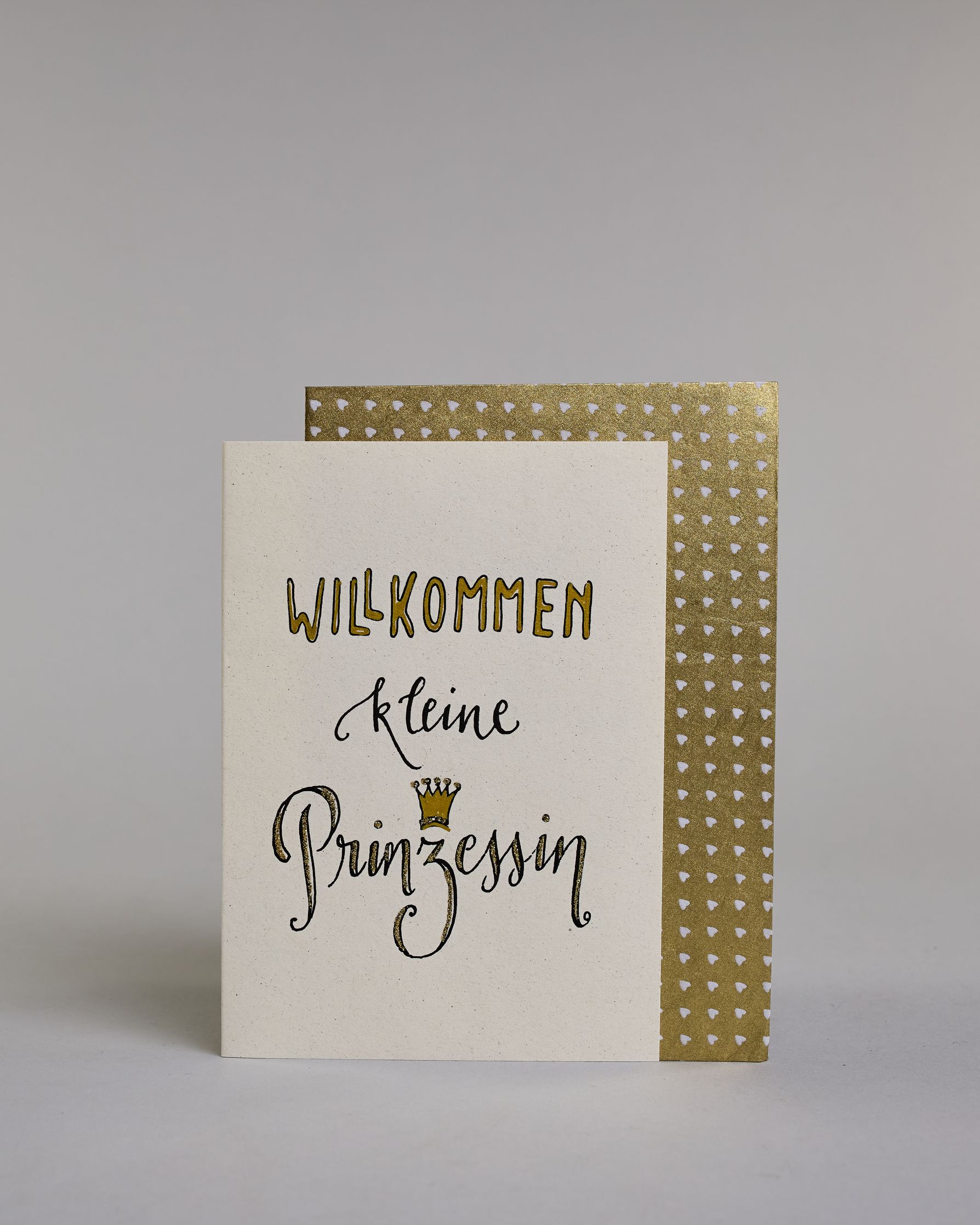 Glückwunschkarte zur Geburt mit schwarzer Kalligrafieaufschrift ‚Willkommen kleine Prinzessin‘, der Text setzt sich auf folgenden Zeilen zusammen:  ‚WILLKOMMEN‘ in gold gefüllten Kalligrafiebuchstaben, darunter‚kleine‘ in geschwungener schwarzer Kalligrafieaufschrift, darunter ‚Prinzessin‘ in verglitzerter Kalligrafieaufschrift, über dem ‚z‘ ist eine gezeichnete goldene Krone auf hellem Hintergrund. Dazu ein gold-weißes handgeschöpftes Kuvert. #2