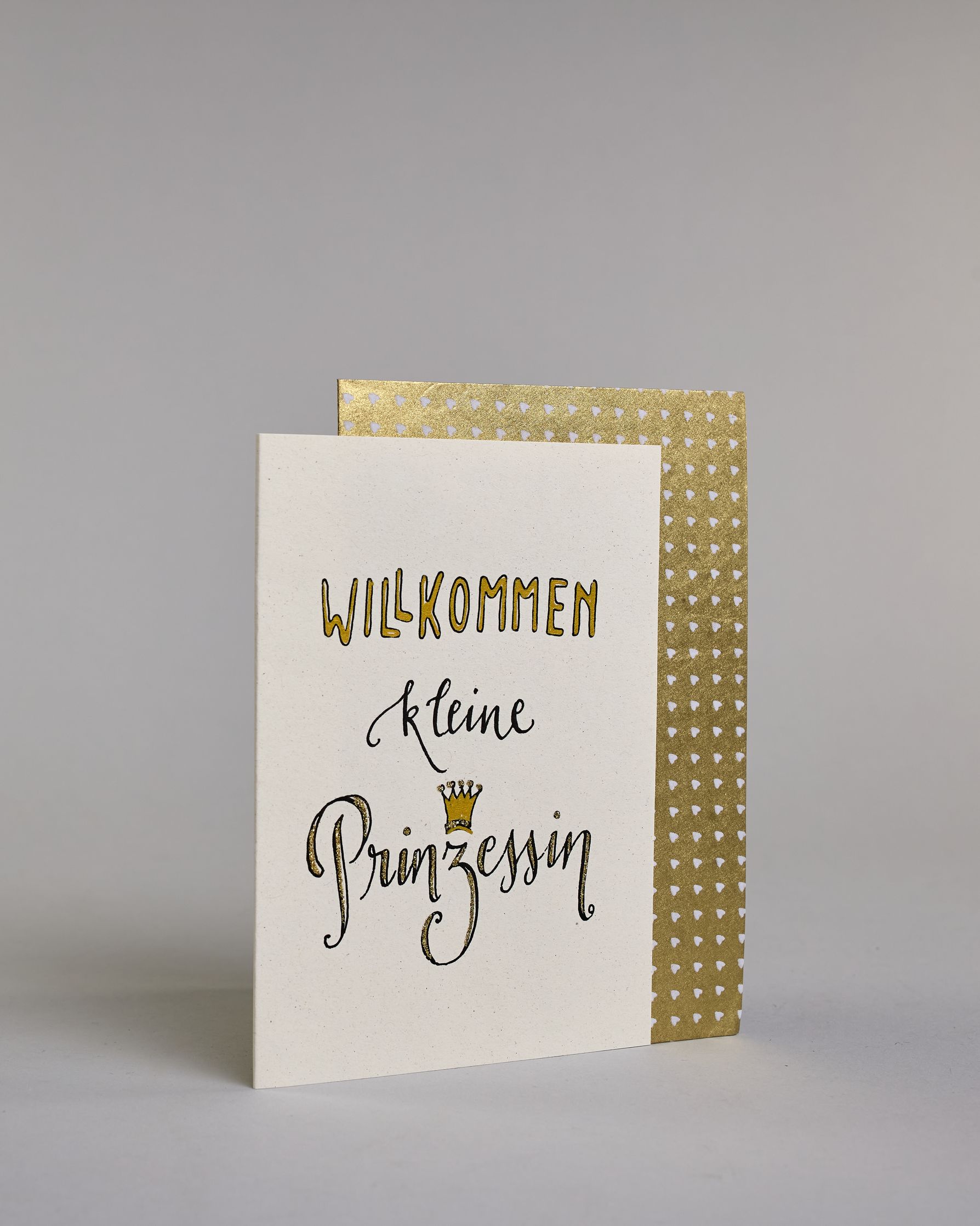 Glückwunschkarte zur Geburt mit schwarzer Kalligrafieaufschrift ‚Willkommen kleine Prinzessin‘, der Text setzt sich auf folgenden Zeilen zusammen:  ‚WILLKOMMEN‘ in gold gefüllten Kalligrafiebuchstaben, darunter‚kleine‘ in geschwungener schwarzer Kalligrafieaufschrift, darunter ‚Prinzessin‘ in verglitzerter Kalligrafieaufschrift, über dem ‚z‘ ist eine gezeichnete goldene Krone auf hellem Hintergrund. Dazu ein gold-weißes handgeschöpftes Kuvert. #3