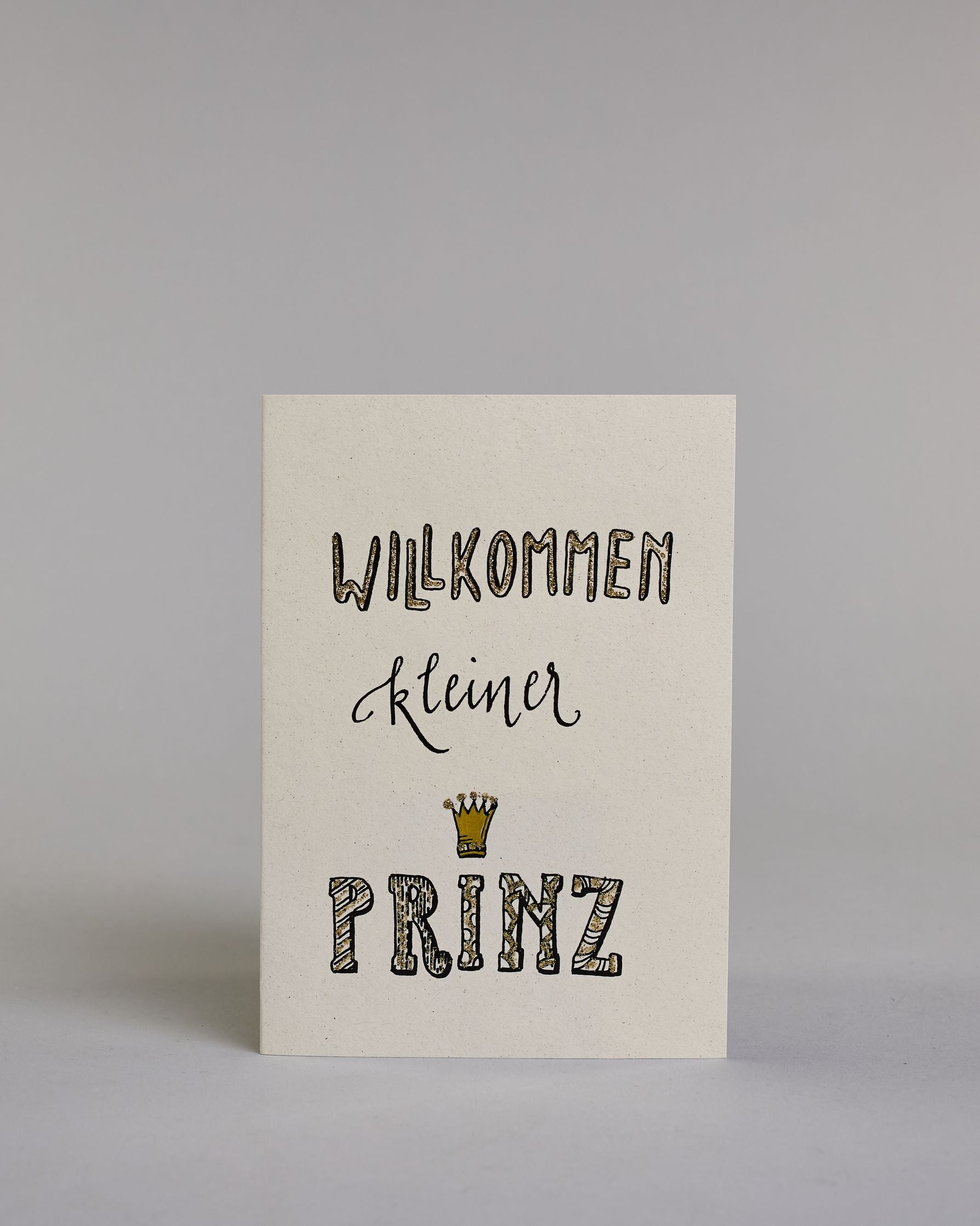 Glückwunschkarte zur Geburt mit der Aufschrift „Willkommen kleiner Prinz“ in schwarzer Kalligrafieaufschrift, wobei das Wort „Willkommen" und „Prinz“ mit goldfarbenen Details verziert ist. Über dem Wort „Prinz“ ist eine kleine goldene Krone abgebildet. Dazu ein grün-goldenes handgeschöpftes Kuvert. #1