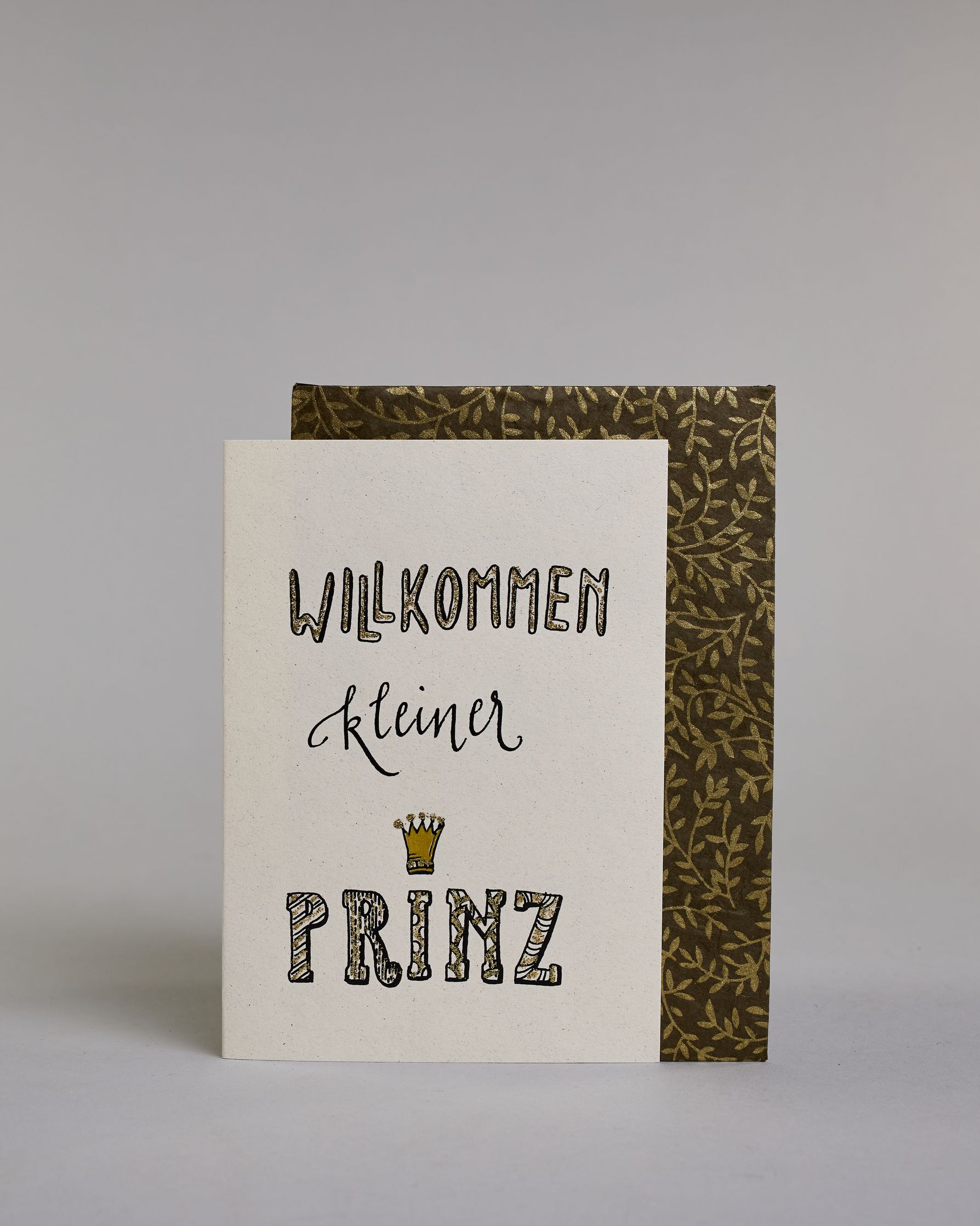 Glückwunschkarte zur Geburt mit der Aufschrift „Willkommen kleiner Prinz“ in schwarzer Kalligrafieaufschrift, wobei das Wort „Willkommen" und „Prinz“ mit goldfarbenen Details verziert ist. Über dem Wort „Prinz“ ist eine kleine goldene Krone abgebildet. Dazu ein grün-goldenes handgeschöpftes Kuvert. #2