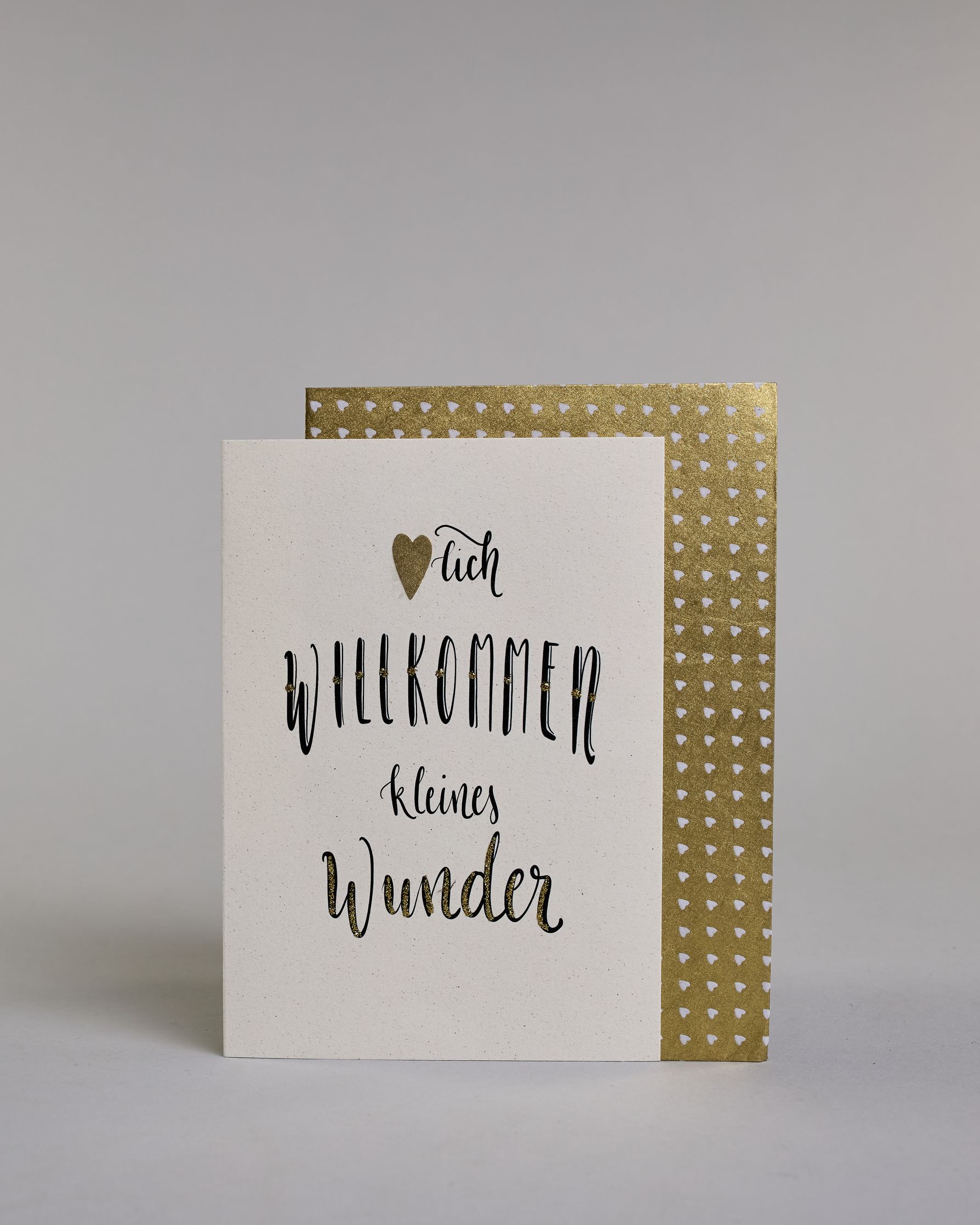 Glückwunschkarte zur Geburt mit der Aufschrift „Herzlich willkommen kleine Wunder“ in schwarzer Kalligrafieaufschrift, wobei in dem Wort „Herzlich" das "Herz" durch ein aufgeklebtes goldenes Herz ersetzt wurde. Das Wort „Wunder“ ist mit goldfarbenen Details verziert. Über dem Wort „Prinz“ ist eine kleine goldene Krone abgebildet. Dazu ein gold-weißes handgeschöpftes Kuvert. #2