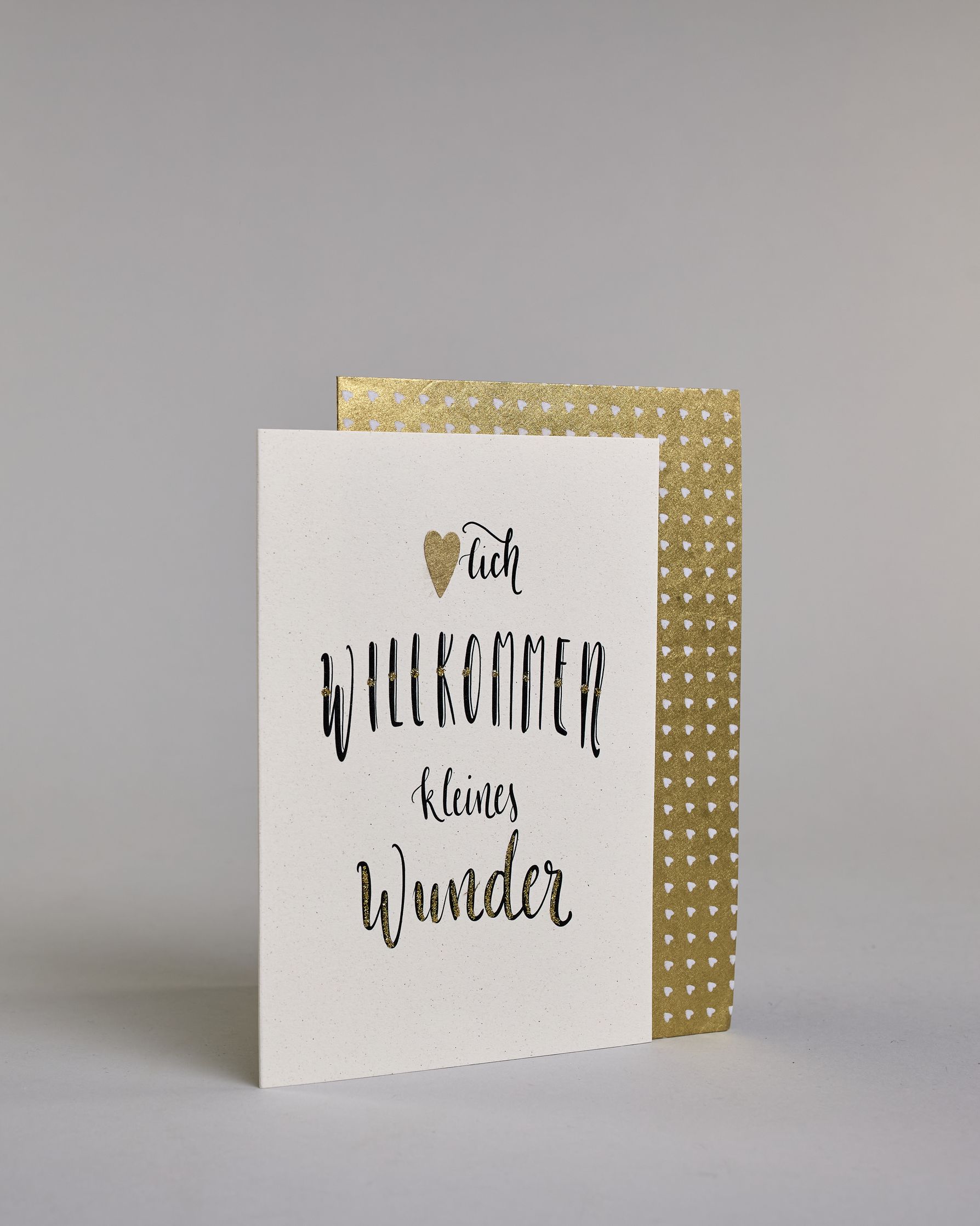 Glückwunschkarte zur Geburt mit der Aufschrift „Herzlich willkommen kleine Wunder“ in schwarzer Kalligrafieaufschrift, wobei in dem Wort „Herzlich" das "Herz" durch ein aufgeklebtes goldenes Herz ersetzt wurde. Das Wort „Wunder“ ist mit goldfarbenen Details verziert. Über dem Wort „Prinz“ ist eine kleine goldene Krone abgebildet. Dazu ein gold-weißes handgeschöpftes Kuvert. #3