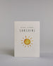 Glückwunschkarte zur Geburt in Englisch mit der Aufschrift „hello little SUNSHINE“ in schwarzer Handlettering-Schrift. Das "hello little" ist in geschwungener Kalligrafieschrift, darunter das"Sunshine" in dünner gerader Schrift. Darunter ist eine gezeichnete gelb-orangefarbene Sonne mit Strahlen. Der Hintergrund ist hell mit kleinen goldenen Punkten. Dazu ein beige-weißes handgeschöpftes Kuvert. #1