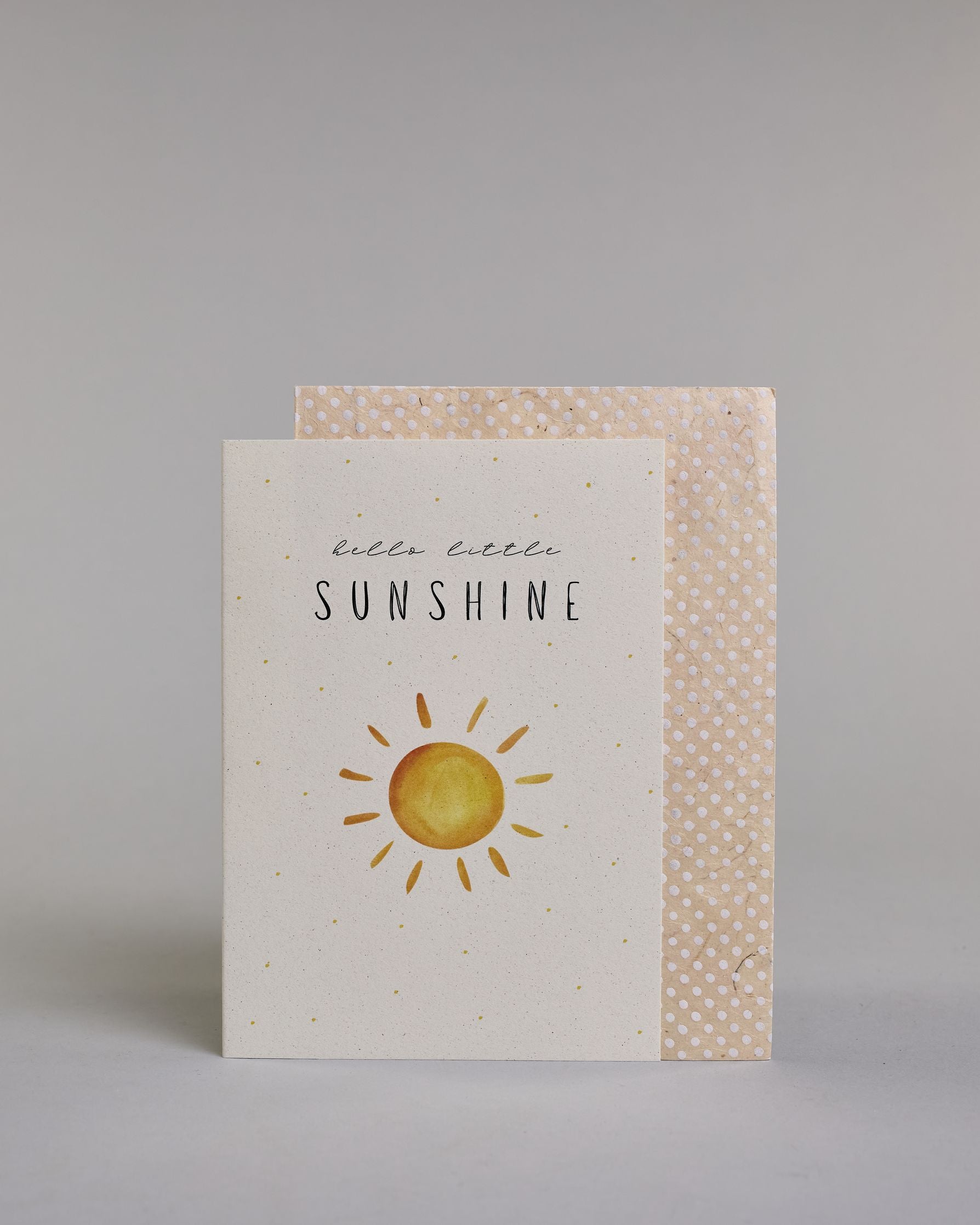 Glückwunschkarte zur Geburt in Englisch mit der Aufschrift „hello little SUNSHINE“ in schwarzer Handlettering-Schrift. Das "hello little" ist in geschwungener Kalligrafieschrift, darunter das"Sunshine" in dünner gerader Schrift. Darunter ist eine gezeichnete gelb-orangefarbene Sonne mit Strahlen. Der Hintergrund ist hell mit kleinen goldenen Punkten. Dazu ein beige-weißes handgeschöpftes Kuvert. #2