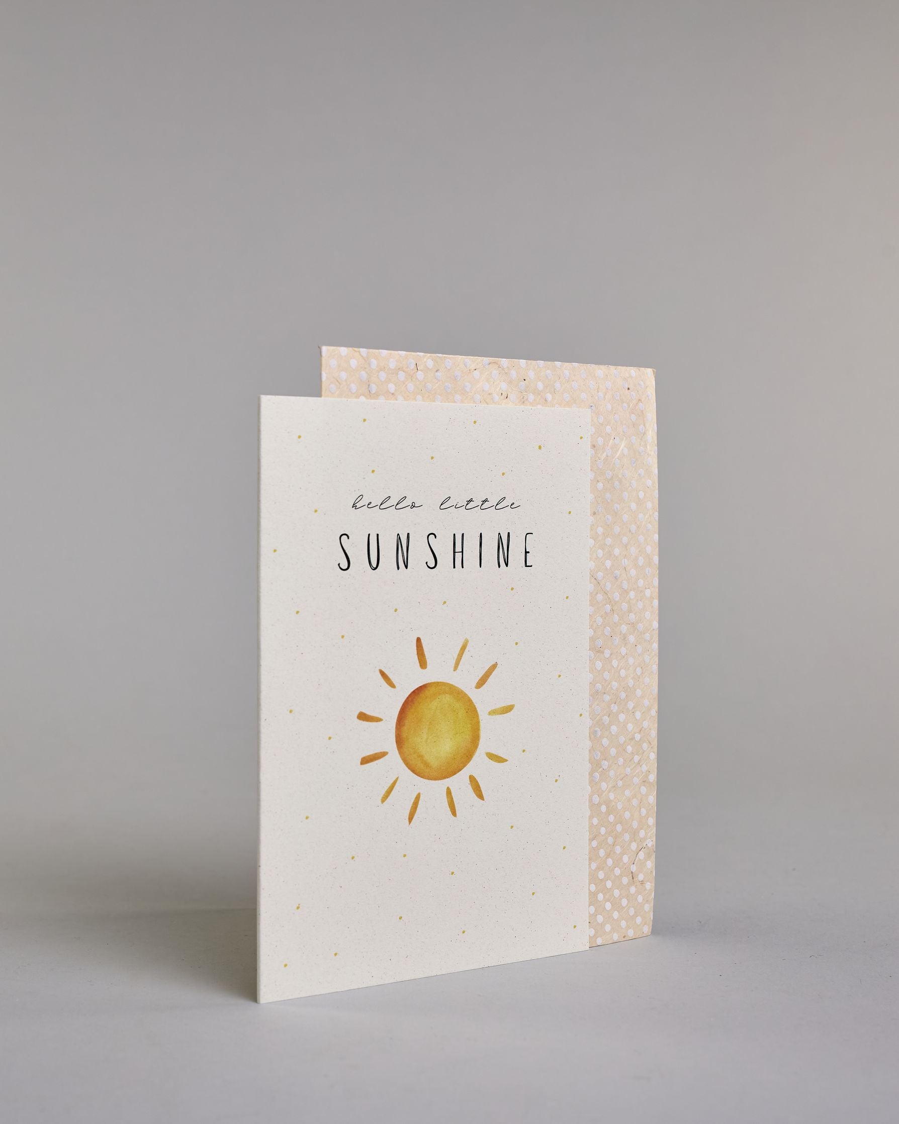 Glückwunschkarte zur Geburt in Englisch mit der Aufschrift „hello little SUNSHINE“ in schwarzer Handlettering-Schrift. Das "hello little" ist in geschwungener Kalligrafieschrift, darunter das"Sunshine" in dünner gerader Schrift. Darunter ist eine gezeichnete gelb-orangefarbene Sonne mit Strahlen. Der Hintergrund ist hell mit kleinen goldenen Punkten. Dazu ein beige-weißes handgeschöpftes Kuvert. #3