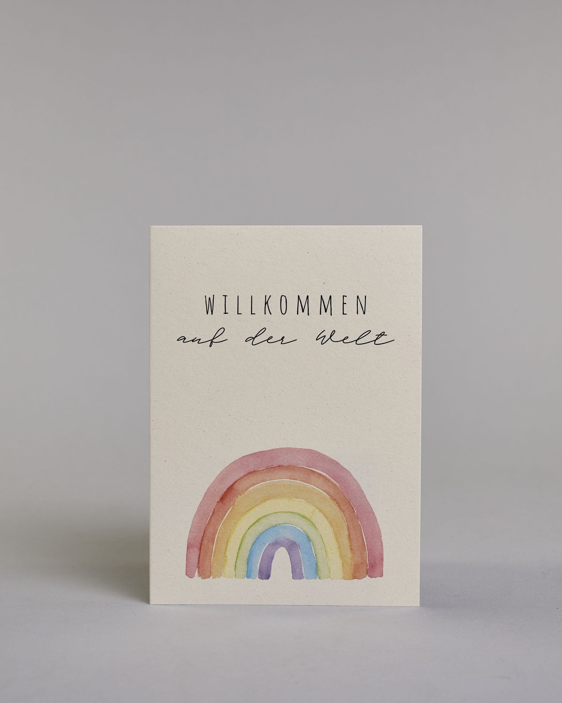 Glückwunschkarte zur Geburt mit der Aufschrift „Willkommen auf der Welt“ in schwarzer Handlettering-Schrift. Darunter ist ein bunter Regenbogen in Aquarelloptik mit den Farben Rot, Orange, Gelb, Grün, Blau und Lila abgebildet. Dazu ein beige-weißes handgeschöpftes Kuvert. #1