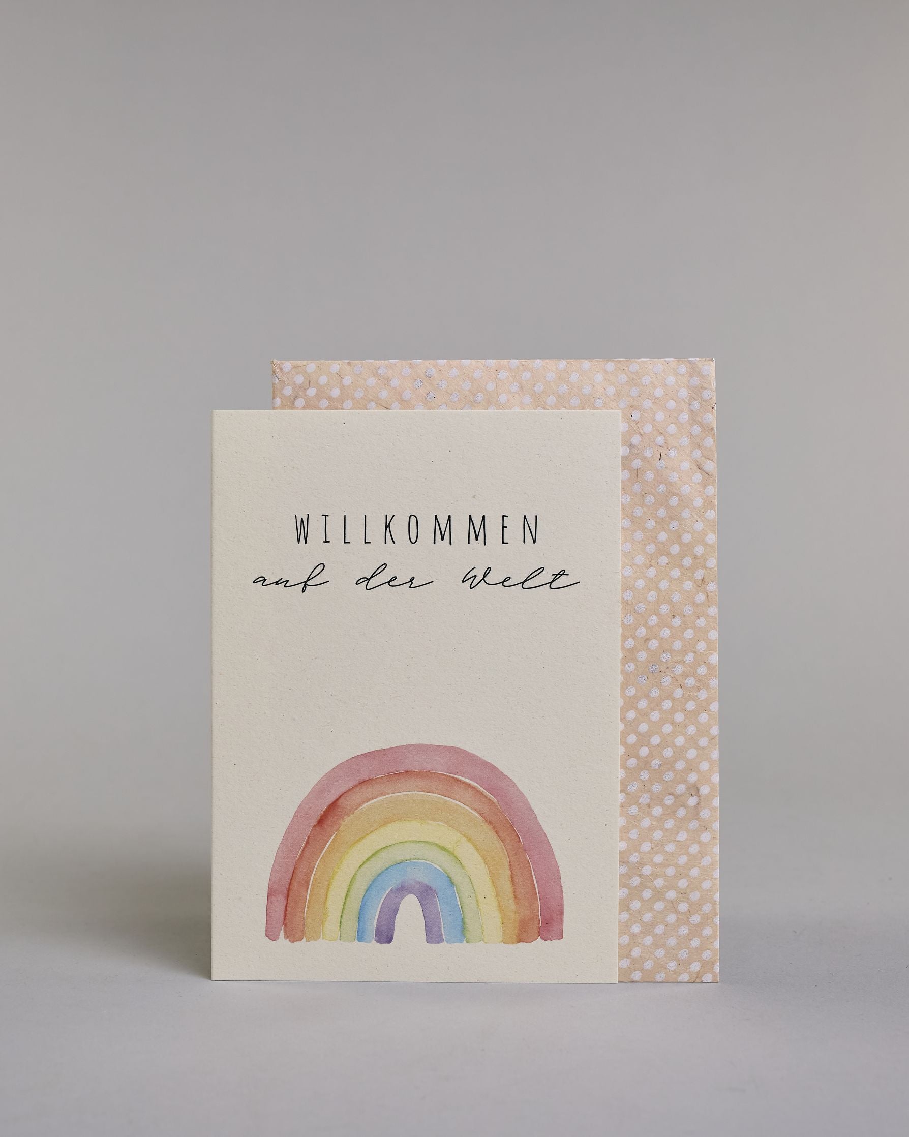 Glückwunschkarte zur Geburt mit der Aufschrift „Willkommen auf der Welt“ in schwarzer Handlettering-Schrift. Darunter ist ein bunter Regenbogen in Aquarelloptik mit den Farben Rot, Orange, Gelb, Grün, Blau und Lila abgebildet. Dazu ein beige-weißes handgeschöpftes Kuvert. #2