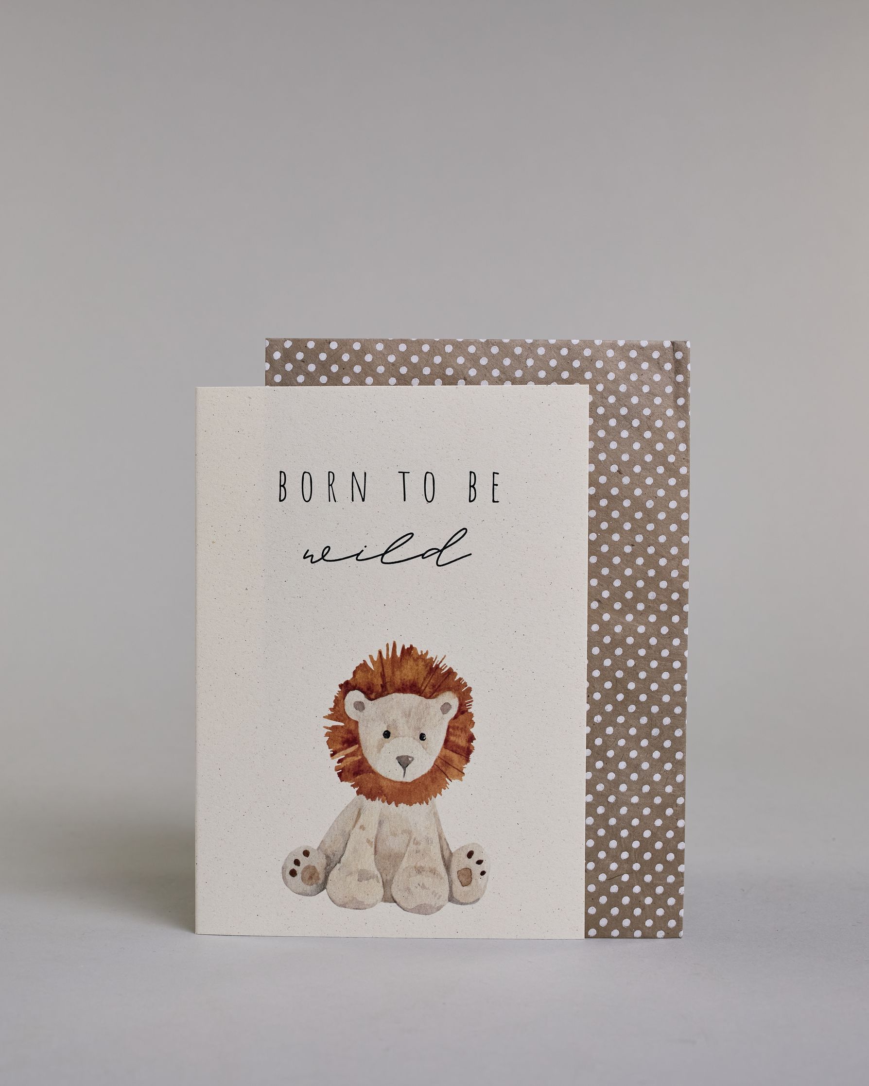 Glückwunschkarte zur Geburt in Englisch mit der Aufschrift „Born to be wild“ in schwarzer Handlettering-Schrift. Das "Born to be" ist in gerader Kalligrafieaufschrift, darunter das "wild" in geschwungener Schrift dargestellt. Darunter ist ein gezeichneter Stofftier-Löwe mit brauner Mähne und hellbeigem Fell abgebildet. Dazu ein beige-weißes handgeschöpftes Kuvert. #2