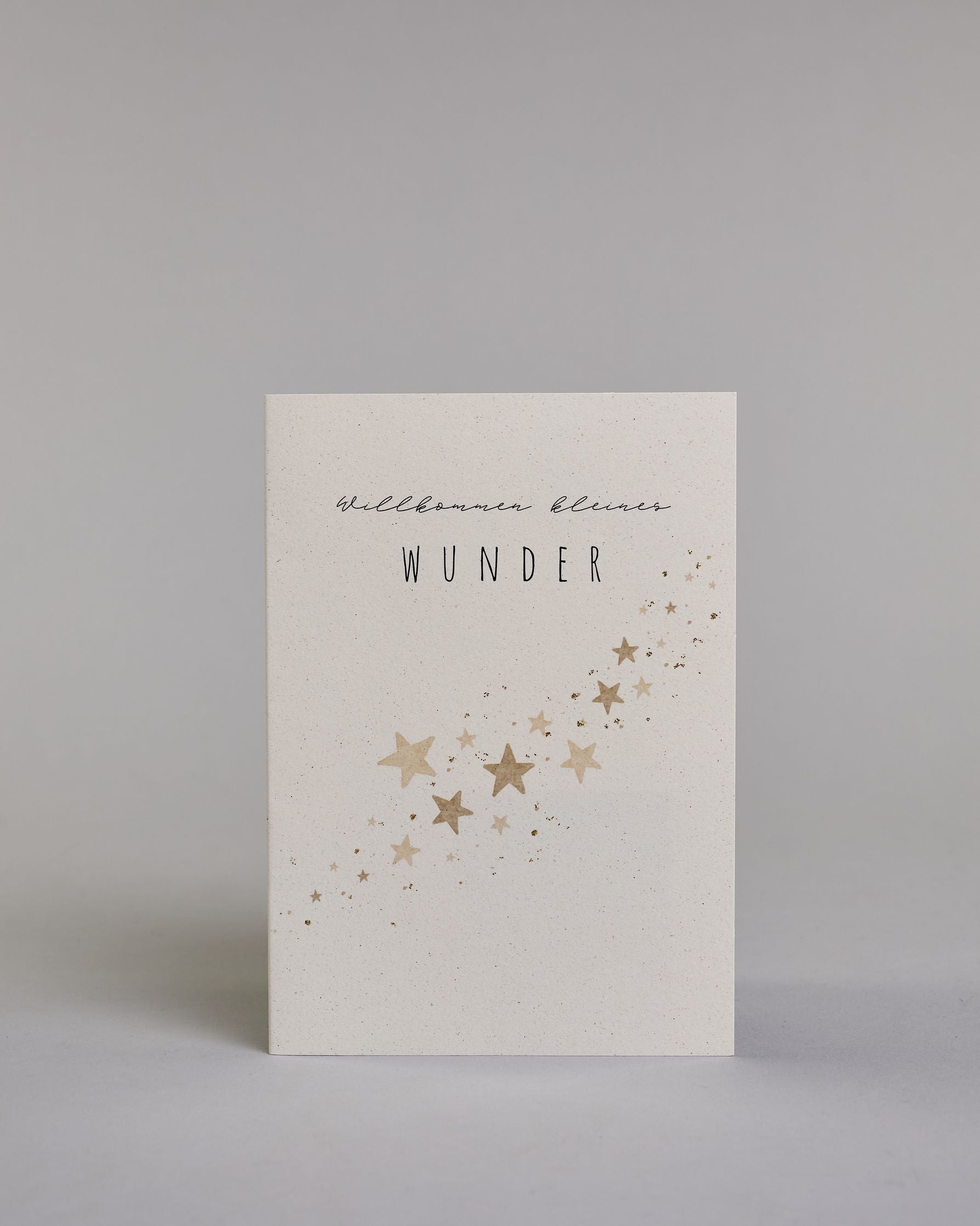Glückwunschkarte zur Geburt mit der Aufschrift „Willkommen kleines Wunder“ in schwarzer Handlettering-Schrift. Das "willkommen kleines" ist in geschwungener Kalligrafieaufschrift, darunter das "WUNDER" in gerader Schrift dargestellt. Darunter verläuft schräg über die Karte ein Schwarm goldfarbener Sterne in verschiedenen Größen. Dazu ein gold-weißes handgeschöpftes Kuvert. #1
