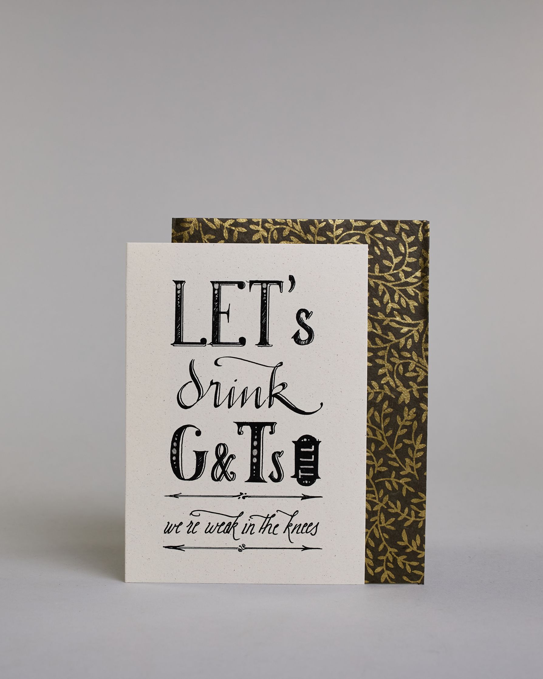 Karte in Englisch mit der Aufschrift „LET’s drink G&Ts till we’re weak in the knees“ in dekorativer schwarzer Handlettering-Schrift auf hellem Hintergrund. Die Zeilen setzten sich wiefolgt zusammen: "LET's" in gerader Schrift, darunter "drink" in geschwungener Schrift, darunte "G&Ts" in gerader Schrift, in der gleichen Zeile rechts daneben vertikal das "till" in einem Rahmen,  darunter "we're weak in the knees". Dazu ein grün-goldenes handgeschöpftes Kuvert. #2