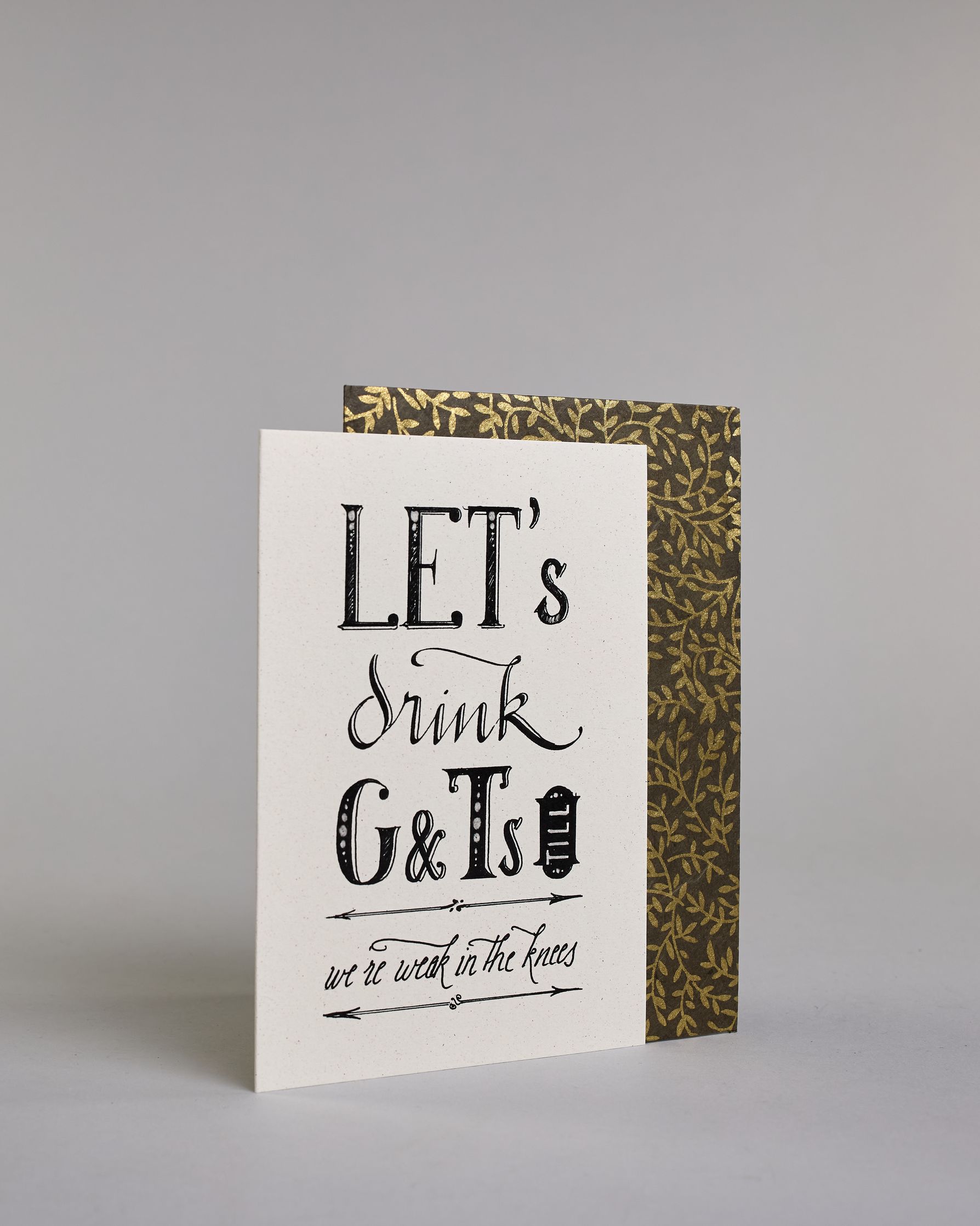 Karte in Englisch mit der Aufschrift „LET’s drink G&Ts till we’re weak in the knees“ in dekorativer schwarzer Handlettering-Schrift auf hellem Hintergrund. Die Zeilen setzten sich wiefolgt zusammen: "LET's" in gerader Schrift, darunter "drink" in geschwungener Schrift, darunte "G&Ts" in gerader Schrift, in der gleichen Zeile rechts daneben vertikal das "till" in einem Rahmen,  darunter "we're weak in the knees". Dazu ein grün-goldenes handgeschöpftes Kuvert. #3