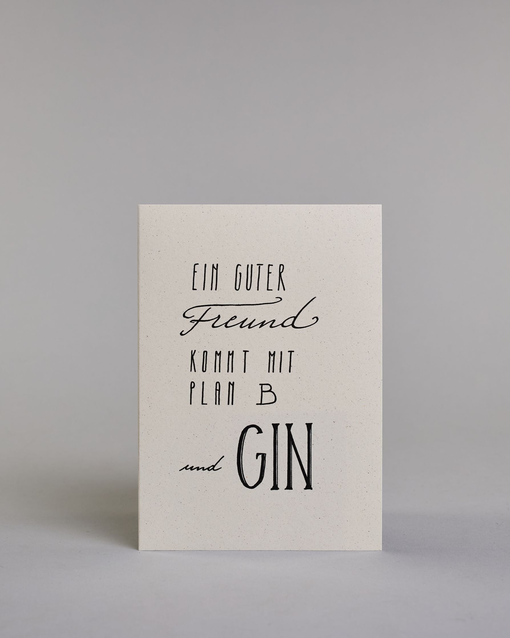 Karte mit der Aufschrift „Ein guter Freund kommt mit Plan B und Gin“ in schwarzer, variierender Handlettering-Schrift auf hellem Hintergrund. Die Zeilen setzten sich wiefolgt zusammen: "Ein guter" in gerader Schrift, darunter "Freund" in geschwungener Schrift, darunter "KOMMT MIT" in gerader Schrift, darunter "PLAN B" in gerader Schrift, darunter "und GIN". Dazu ein grün-goldenes handgeschöpftes Kuvert. #1