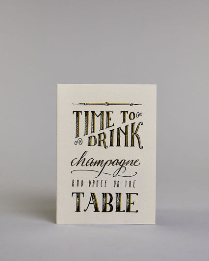 Karte in Englisch mit der Aufschrift „Time to drink champagne and dance on the table“ in dekorativer Handlettering-Schrift auf hellem Hintergrund. Die Wörter sind in Schwarz gestaltet, teils mit goldfarbenen und glitzernden Details hervorgehoben. Das "TIME TO DRINK" und "TABLE" ist in kreativer gerader Kalligrafieaufschrift gestaltet, das "Champagne" in geschwungener und das "AND DANCE ON THE" in dünner gerader Schrift. Dazu ein grün-goldenes handgeschöpftes Kuvert. #1