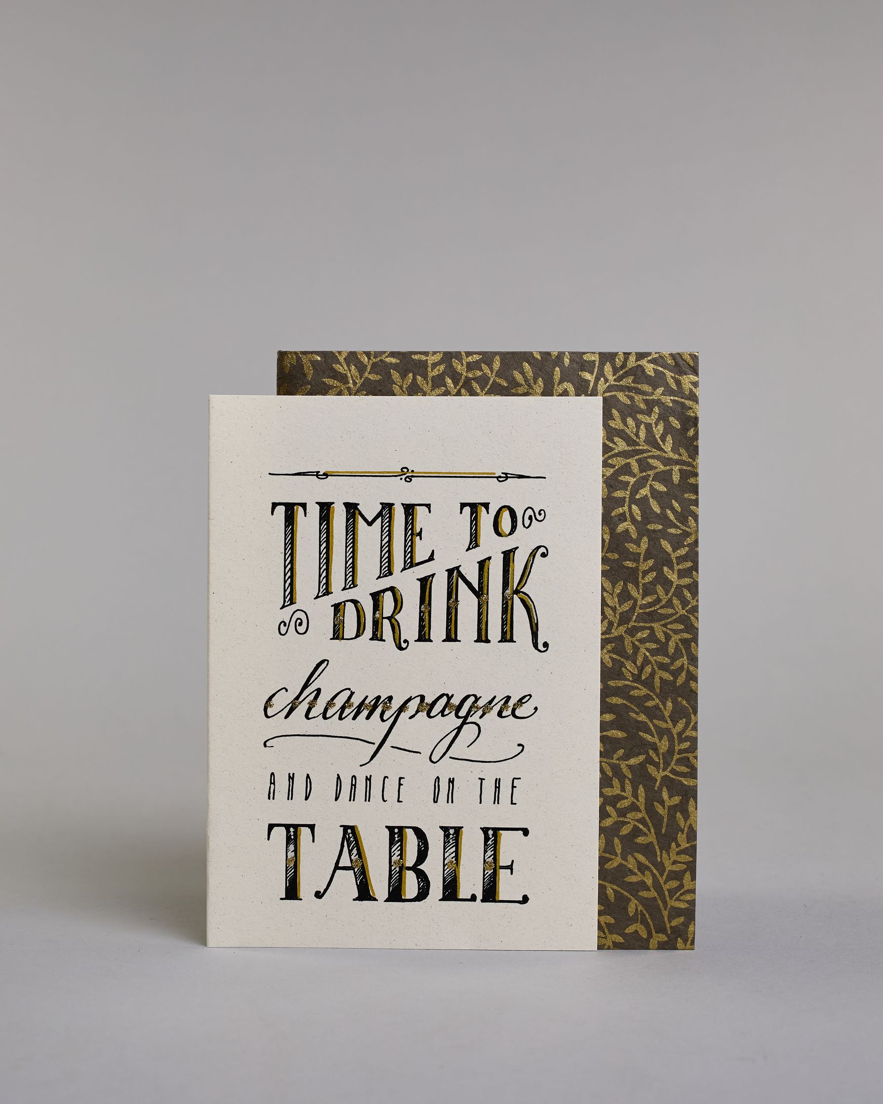 Karte in Englisch mit der Aufschrift „Time to drink champagne and dance on the table“ in dekorativer Handlettering-Schrift auf hellem Hintergrund. Die Wörter sind in Schwarz gestaltet, teils mit goldfarbenen und glitzernden Details hervorgehoben. Das "TIME TO DRINK" und "TABLE" ist in kreativer gerader Kalligrafieaufschrift gestaltet, das "Champagne" in geschwungener und das "AND DANCE ON THE" in dünner gerader Schrift. Dazu ein grün-goldenes handgeschöpftes Kuvert. #2